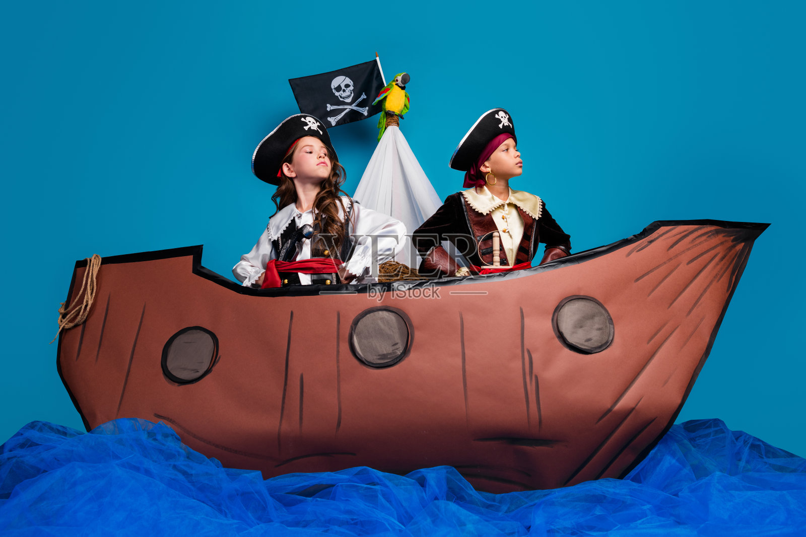 全身照,两个迷人的小孩勇敢自信地摆姿势,穿着海盗万圣节服装,船只装饰,背景为蓝色。照片摄影图片