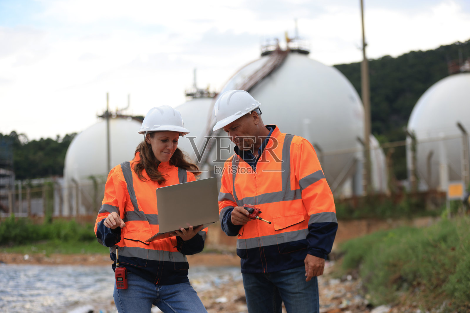 穿着安全工作服的男性和女性高级工程师正在讨论石油和天然气生产性能以及废水研究,使用一台笔记本电脑。照片摄影图片