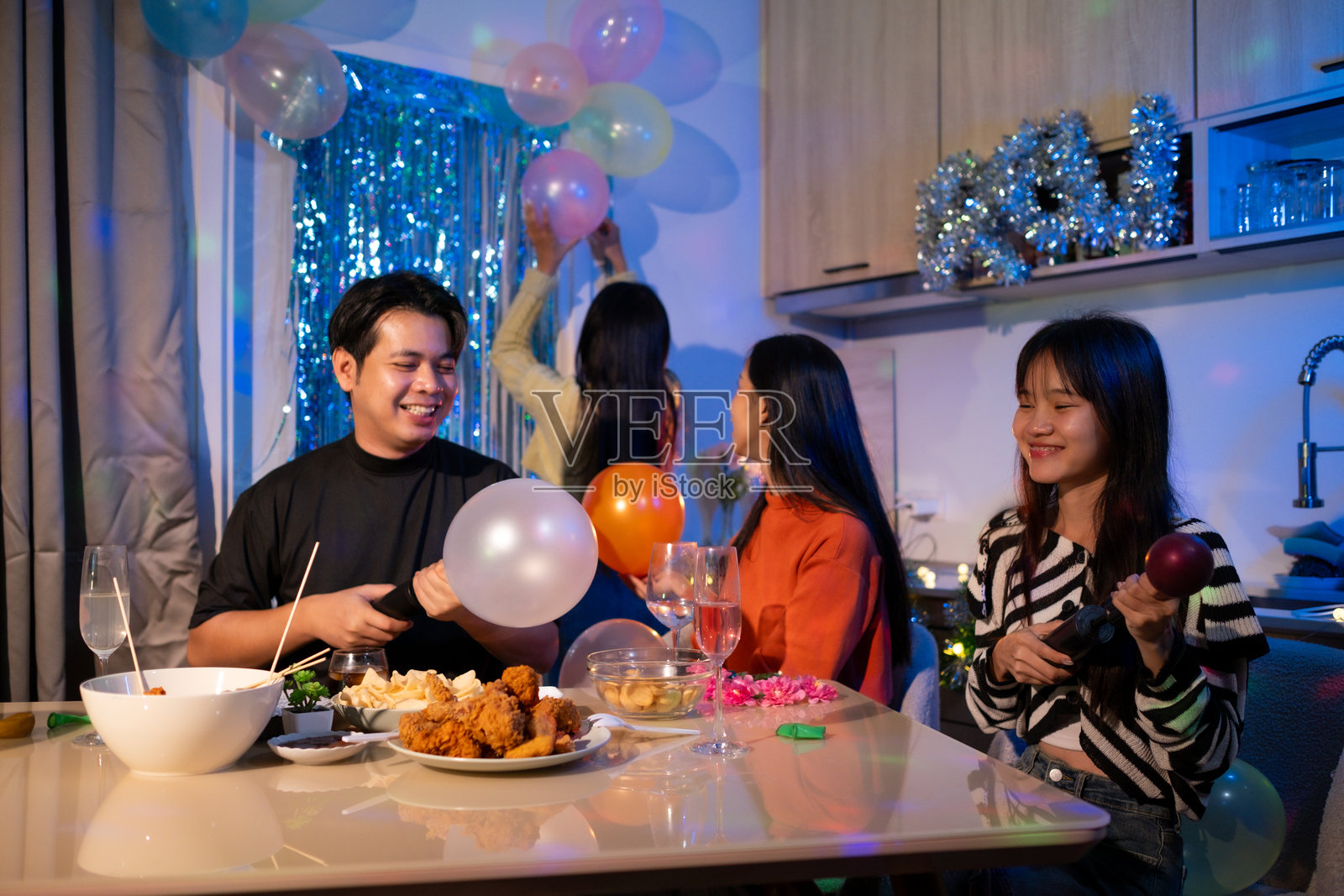 快乐的朋友们在家派对上吹气球,享用小吃和饮料。照片摄影图片