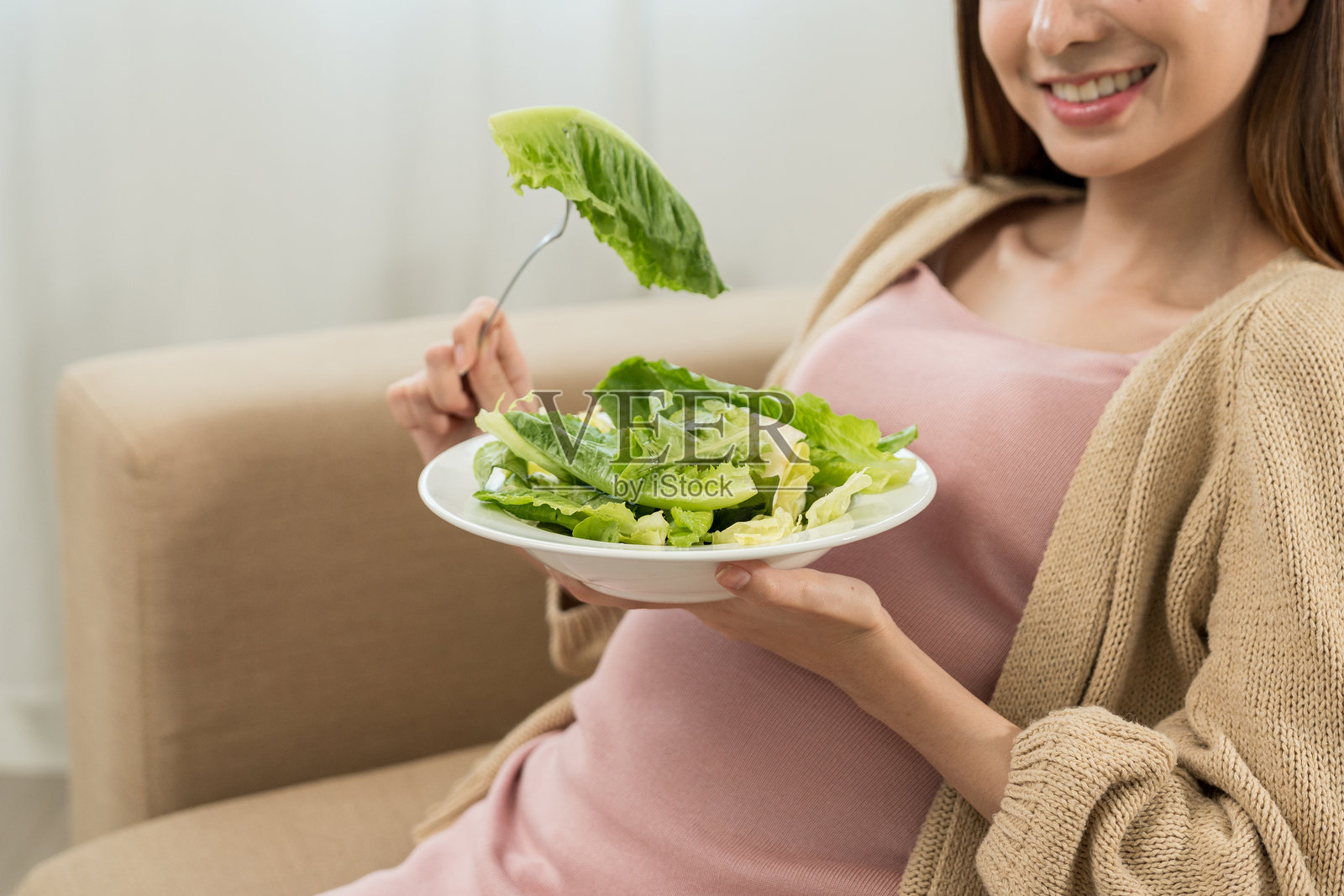 快乐的孕妇吃着蔬菜沙拉,微笑着看向镜头,体现了健康母亲关注自身健康、滋养胎儿并期待分娩的概念。照片摄影图片