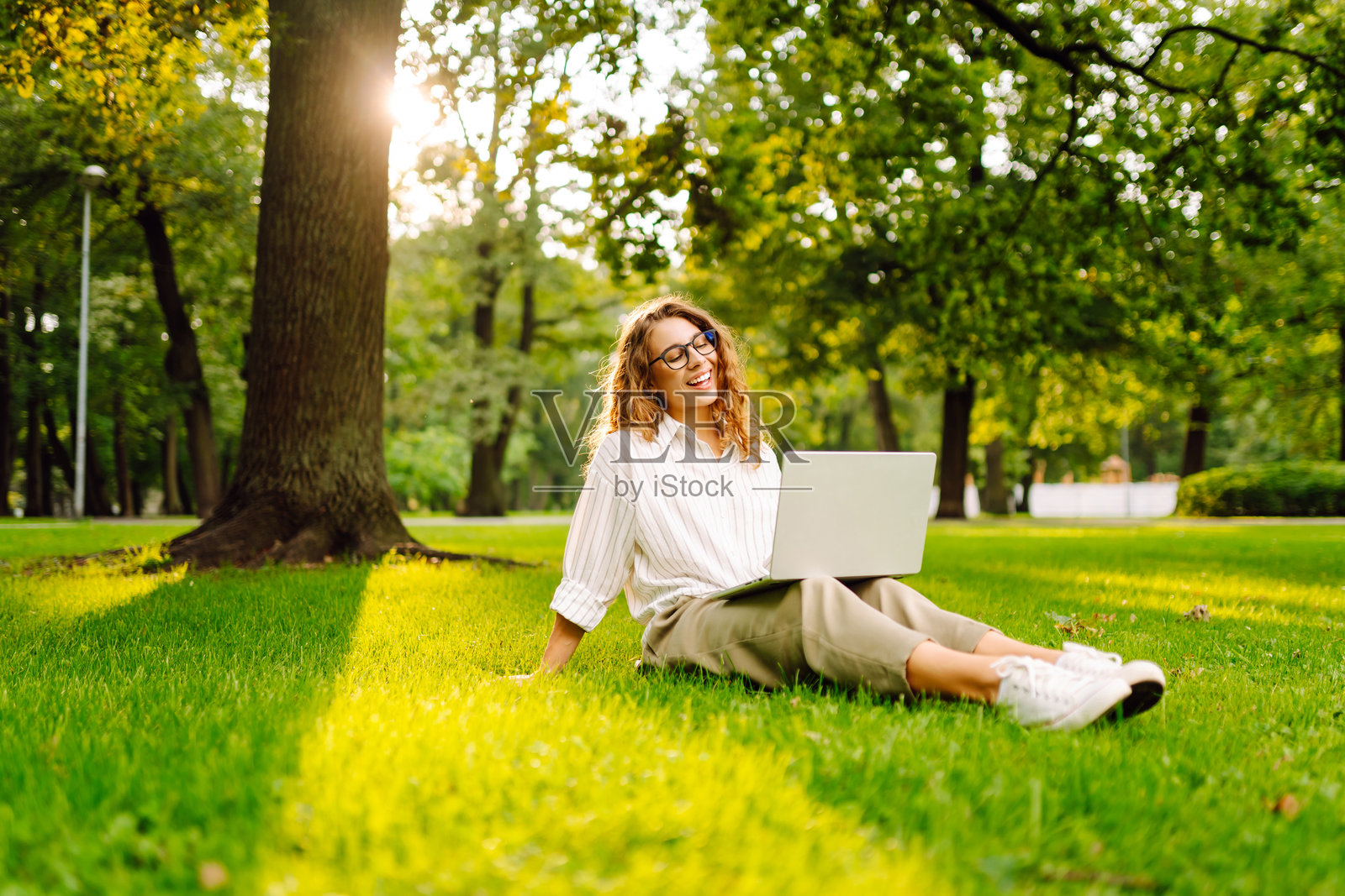 年轻女性坐在城市公园的绿草地上,使用笔记本电脑工作。照片摄影图片
