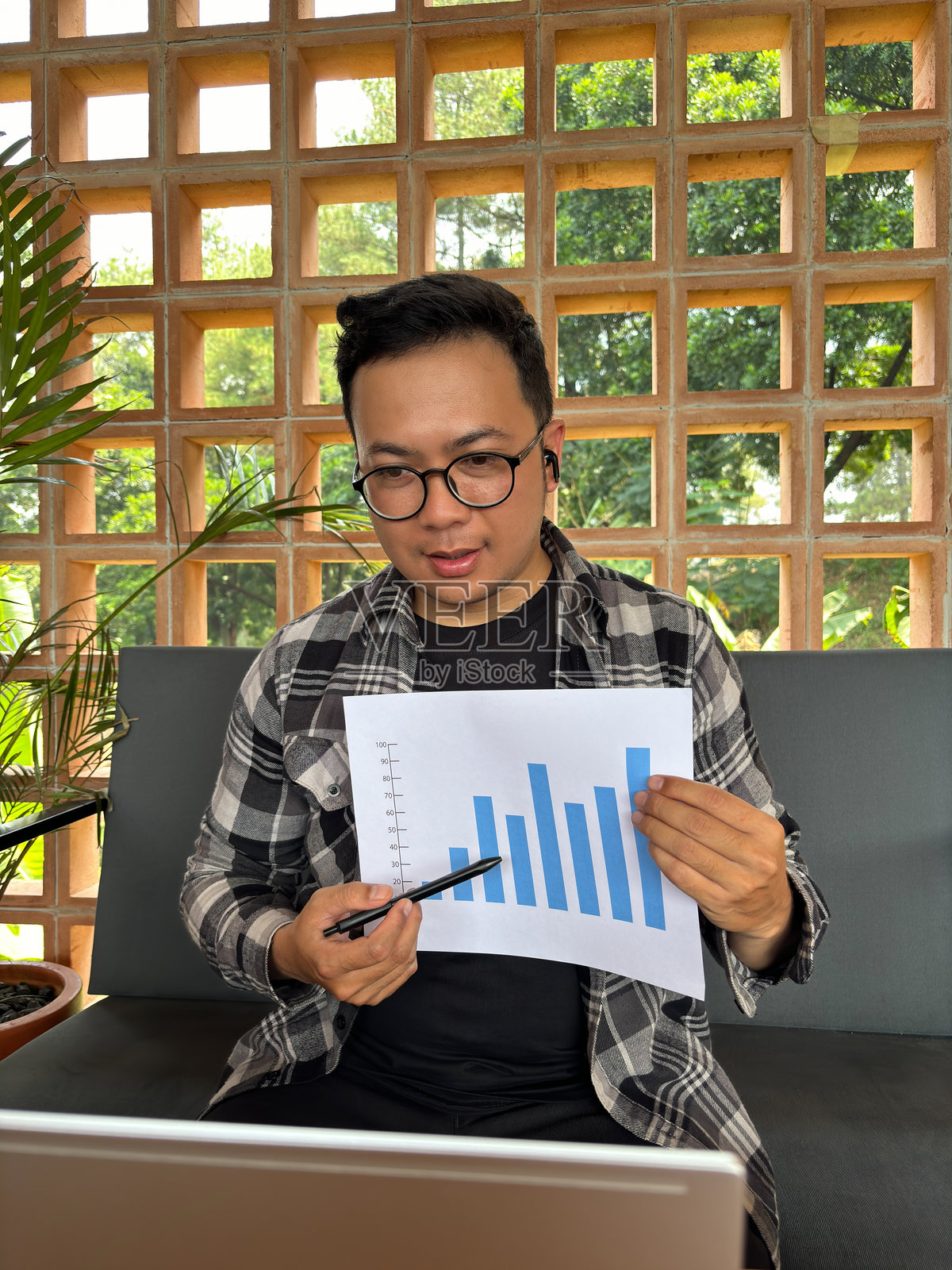 男子在在线会议中使用笔记本电脑,同时手持销售图表文件并用铅笔指着。照片摄影图片