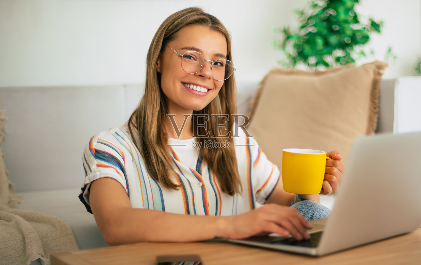 戴眼镜的快乐女性坐在桌子前,正在使用笔记本电脑,手里拿着一个黄色的杯子。她穿着条纹衬衫,打扮休闲,身处一个舒适的家庭环境,非常适合居家办公的主题。照片摄影图片