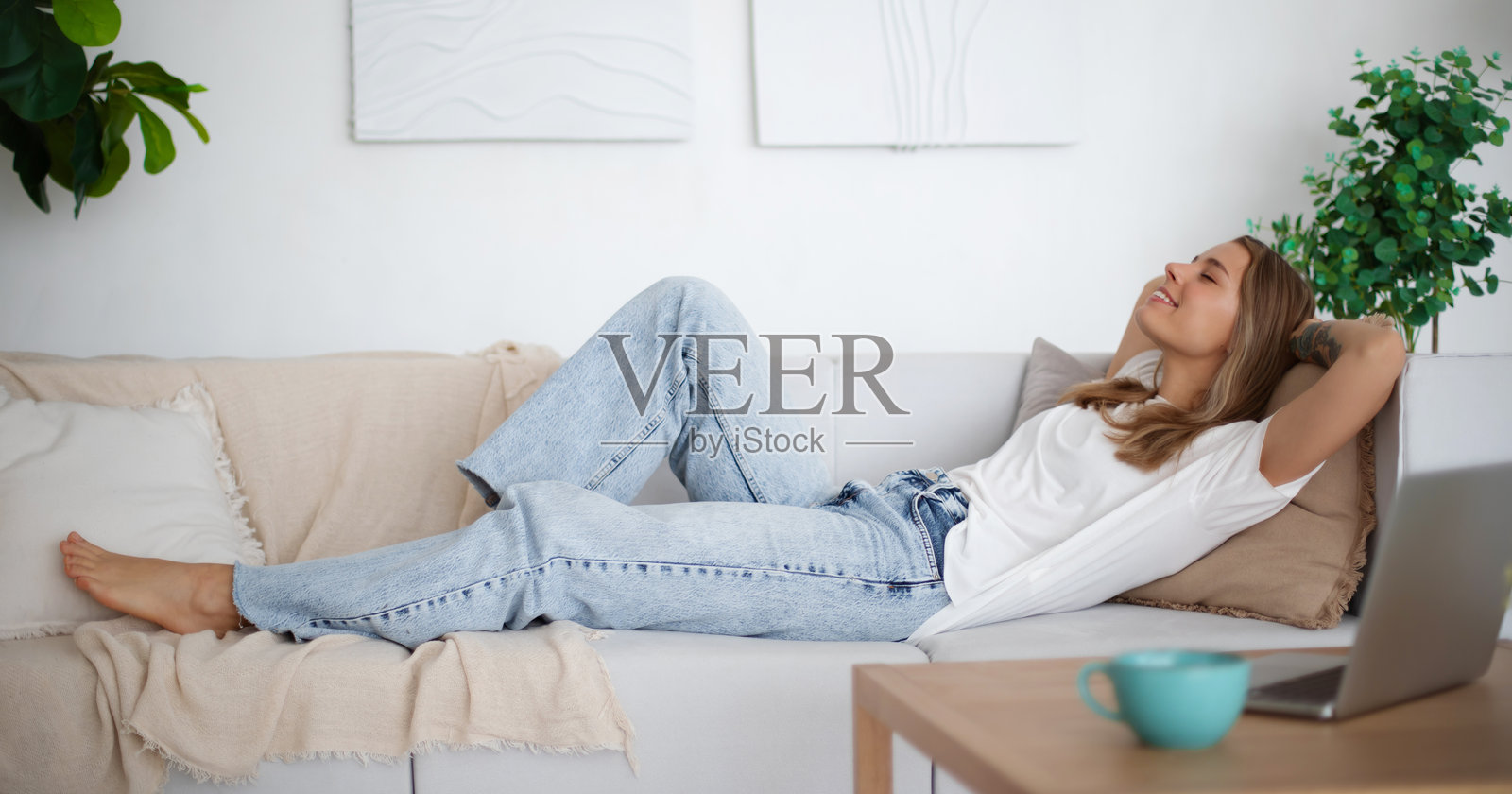 一位年轻女性躺在白色沙发上,双手放在头后,微笑着放松。她穿着休闲的白色T恤和牛仔裤,坐在一个明亮现代的客厅里,周围有柔软的抱枕和绿色植物。照片摄影图片