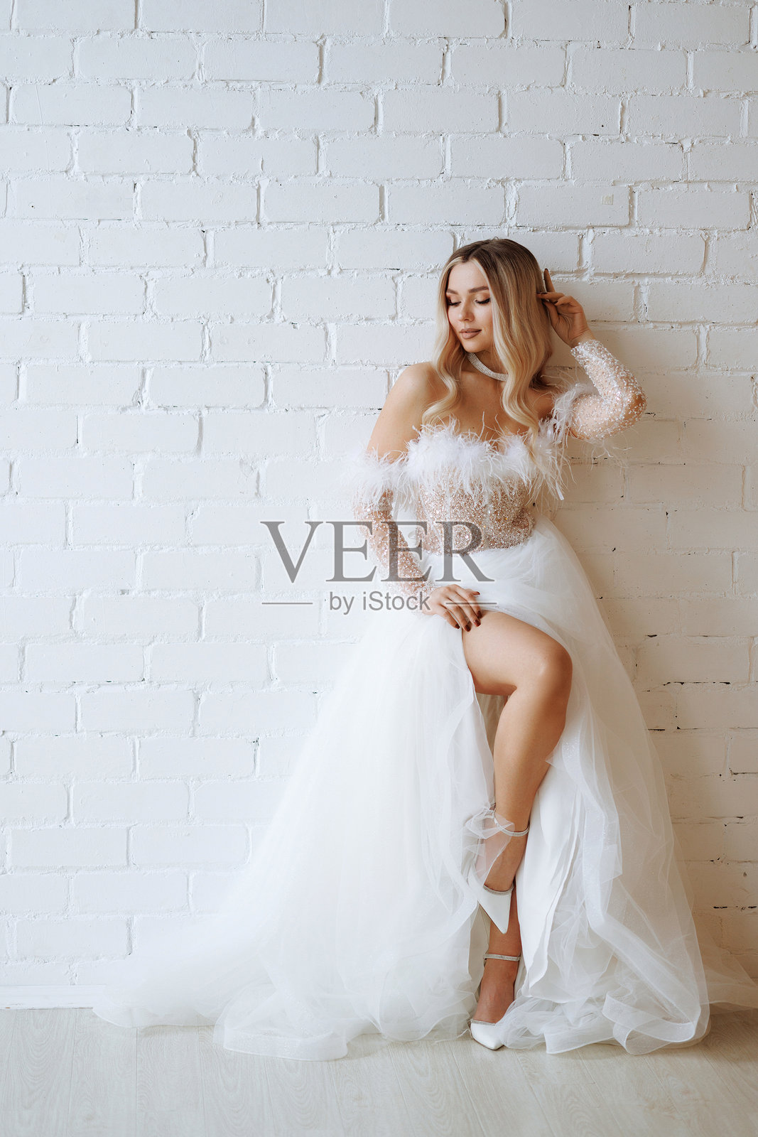 金发新娘穿着白色蓬松婚纱,靠在白色砖墙前摆姿势。照片摄影图片