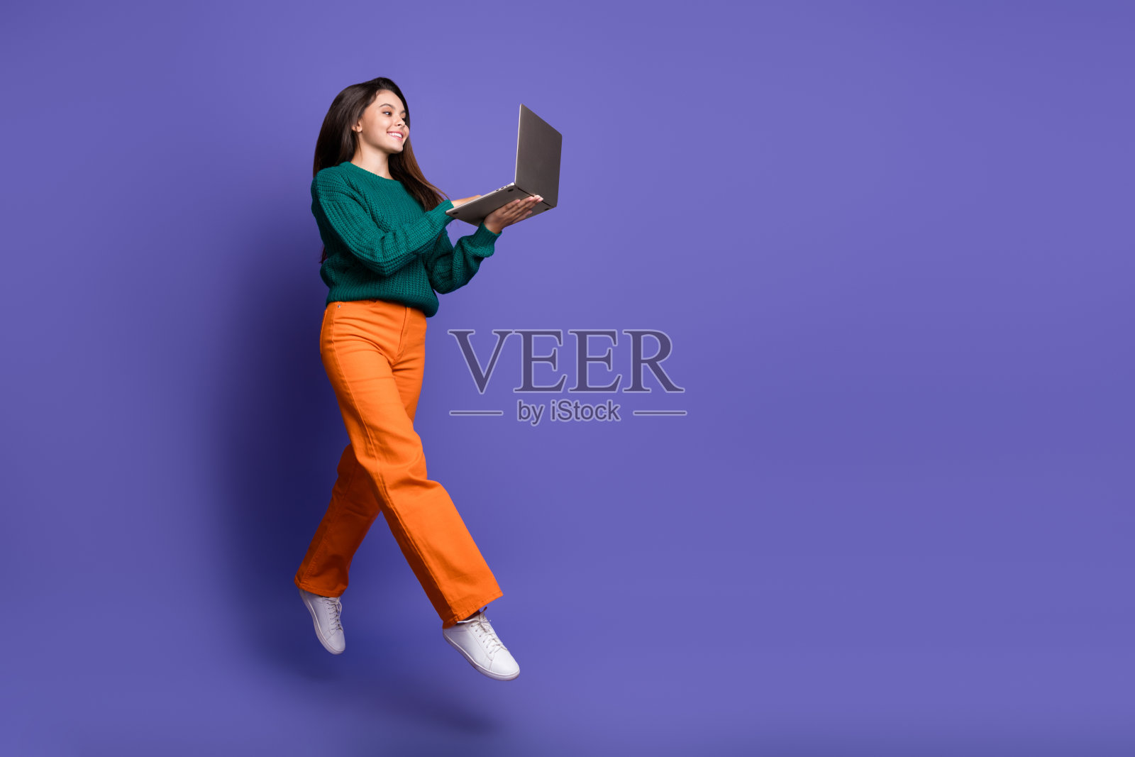 年轻女孩在紫色背景下跳跃,手中拿着笔记本电脑,展现出活力四射的全身肖像。照片摄影图片