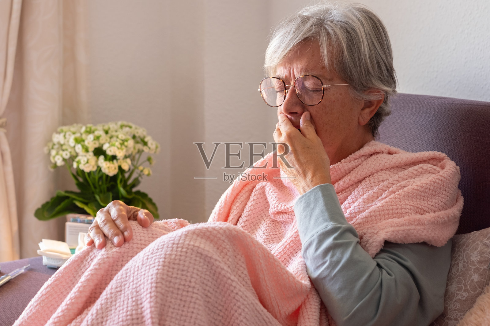 患有严重感冒和发烧症状的白人老年女性,可能是季节性流感、COVID或花粉过敏 - 她在家沙发上自我用药,身边有药片、温度计和毯子。照片摄影图片