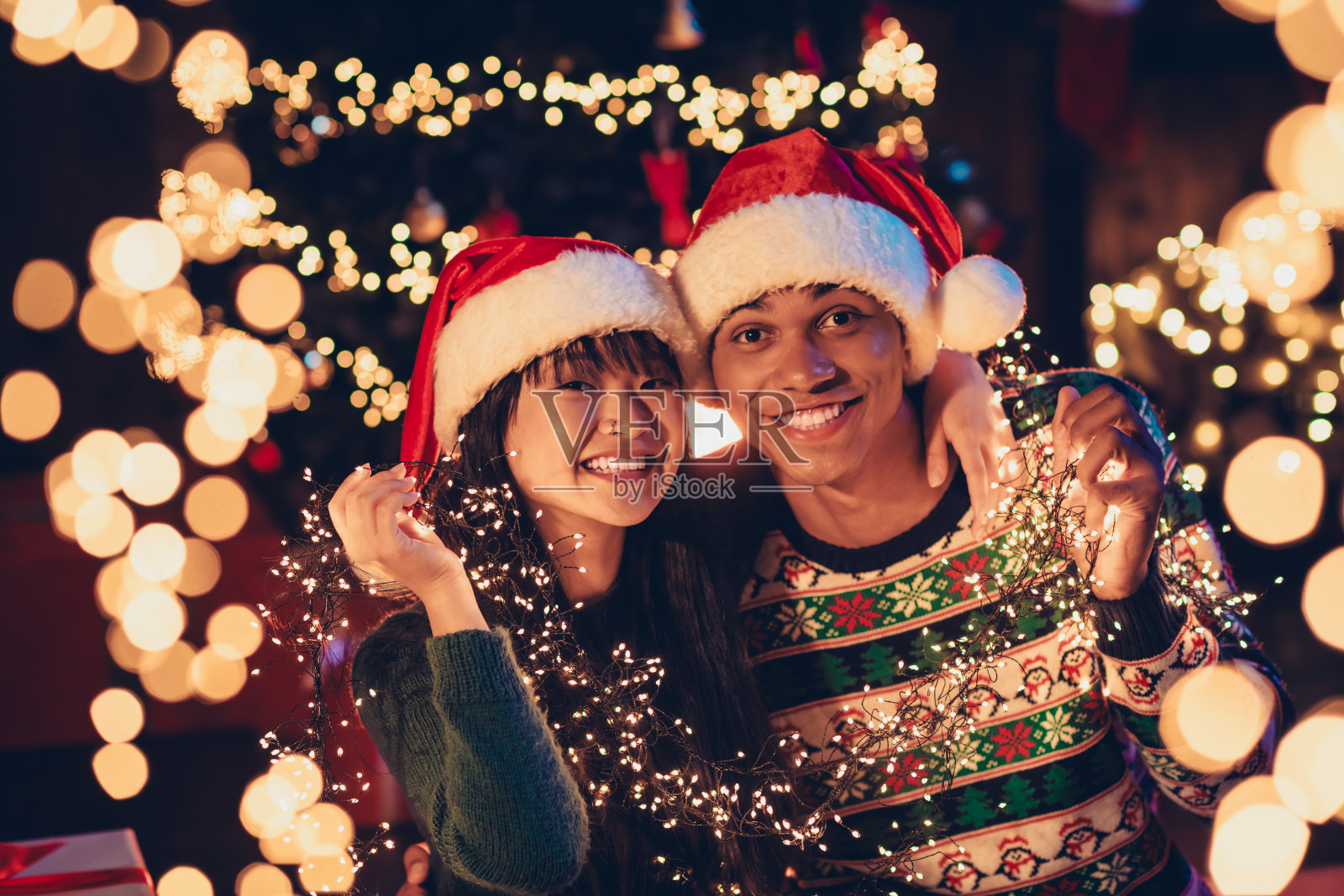 一对年轻情侣在室内拥抱庆祝圣诞新年,手持灯饰花环。照片摄影图片