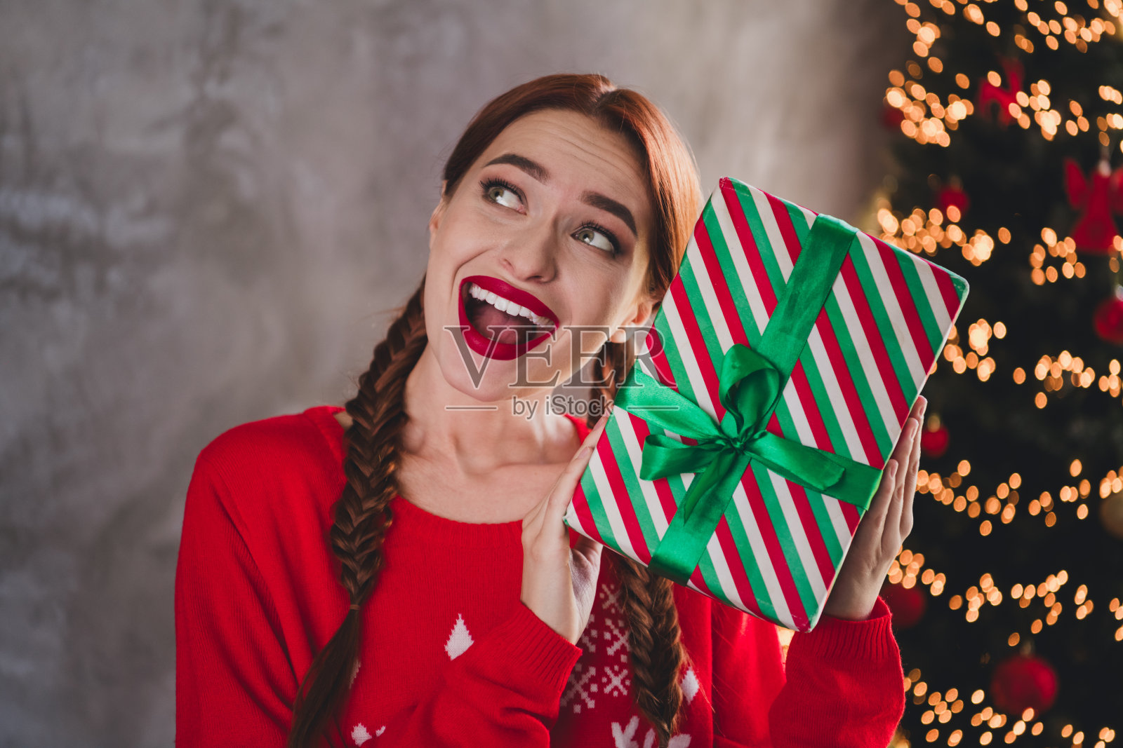 可爱的年轻红发女性兴奋地拿着礼物,穿着圣诞服装,室内派对装饰,阳光明媚的家居环境。照片摄影图片