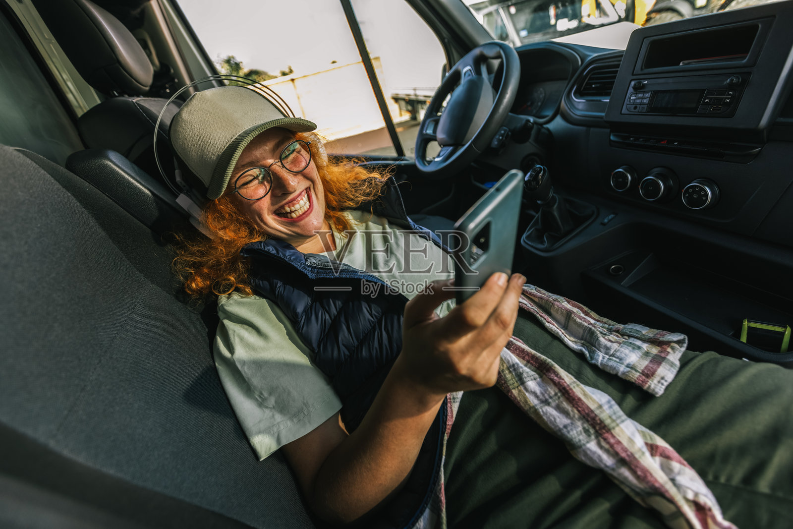 年轻的红发女卡车司机在休息,使用手机和耳机听音乐。照片摄影图片