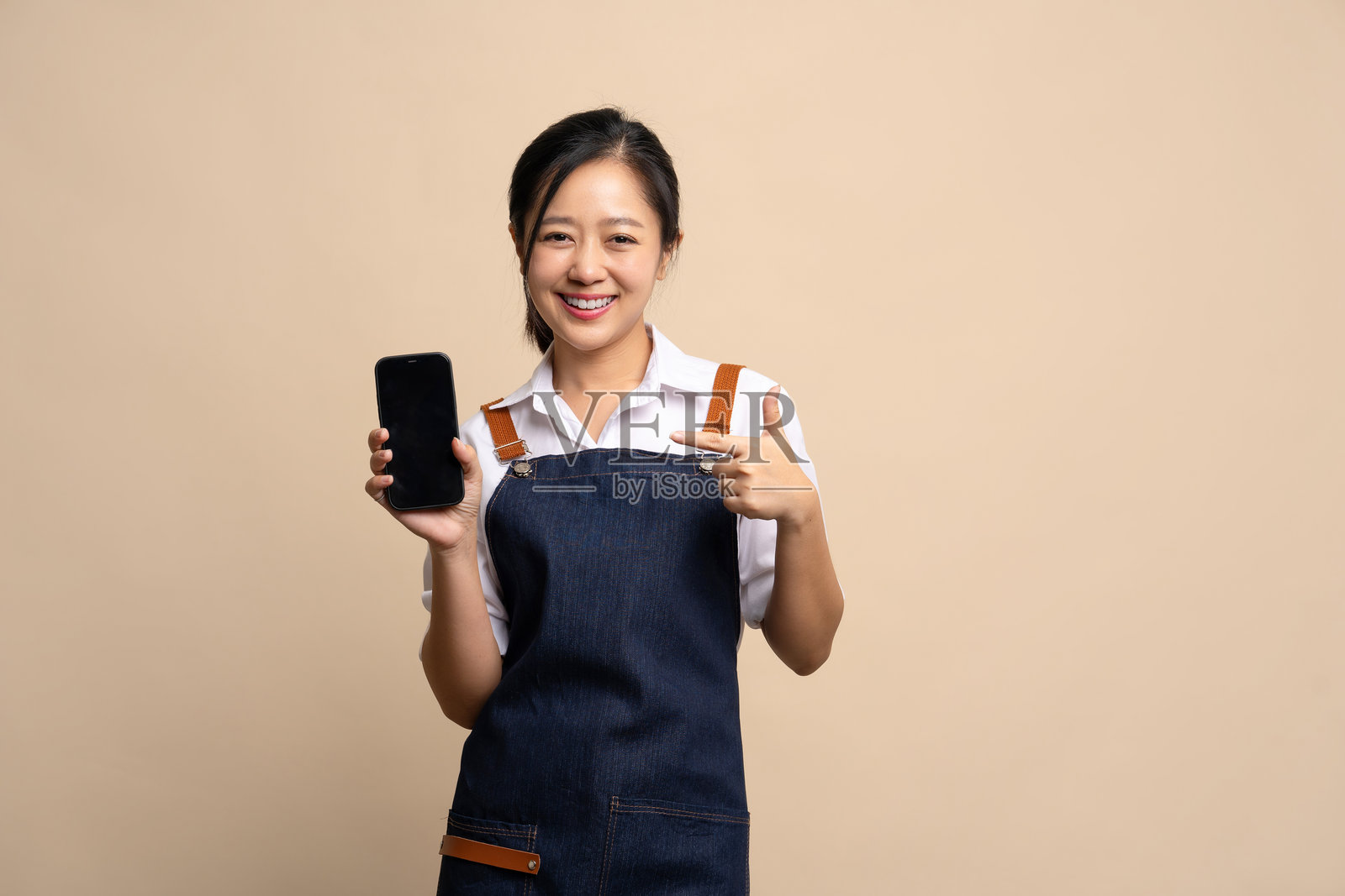 微笑的年轻亚洲女性穿着围裙,站在米色背景前。她手持智能手机,用手指指向空白屏幕。照片摄影图片