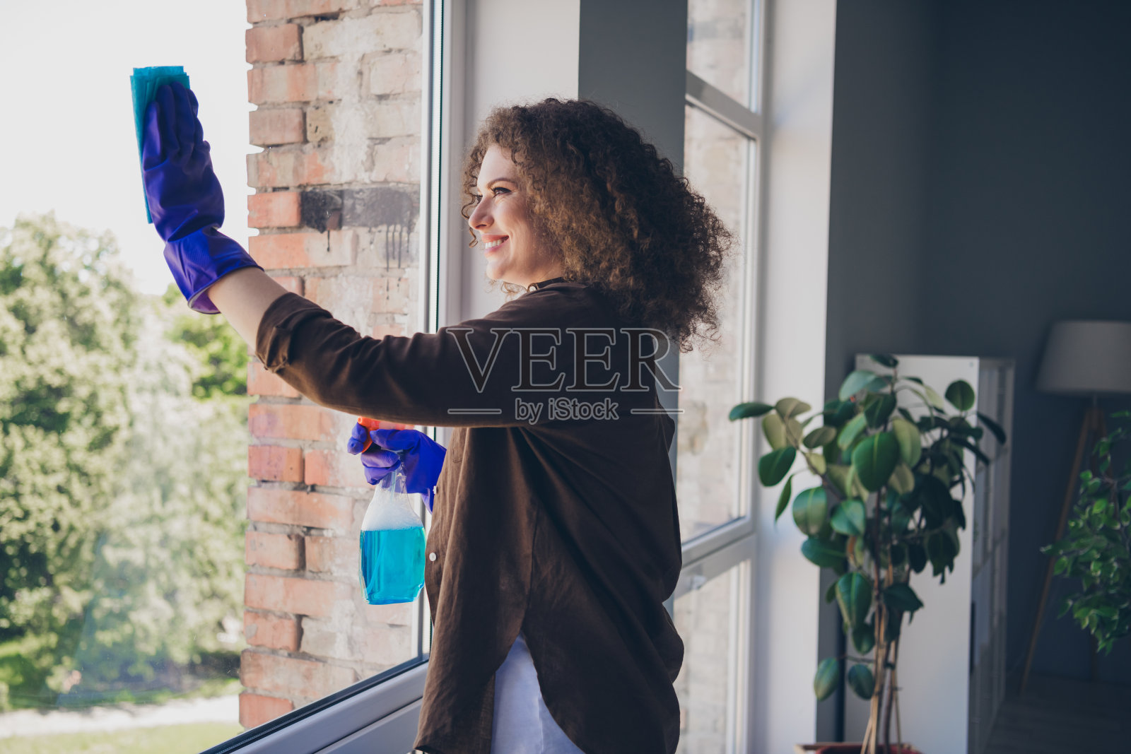 年轻的家庭主妇穿着休闲衬衫,使用柔软的抹布在舒适的阁楼公寓内擦拭巨大的窗户,使用清洁剂。照片摄影图片