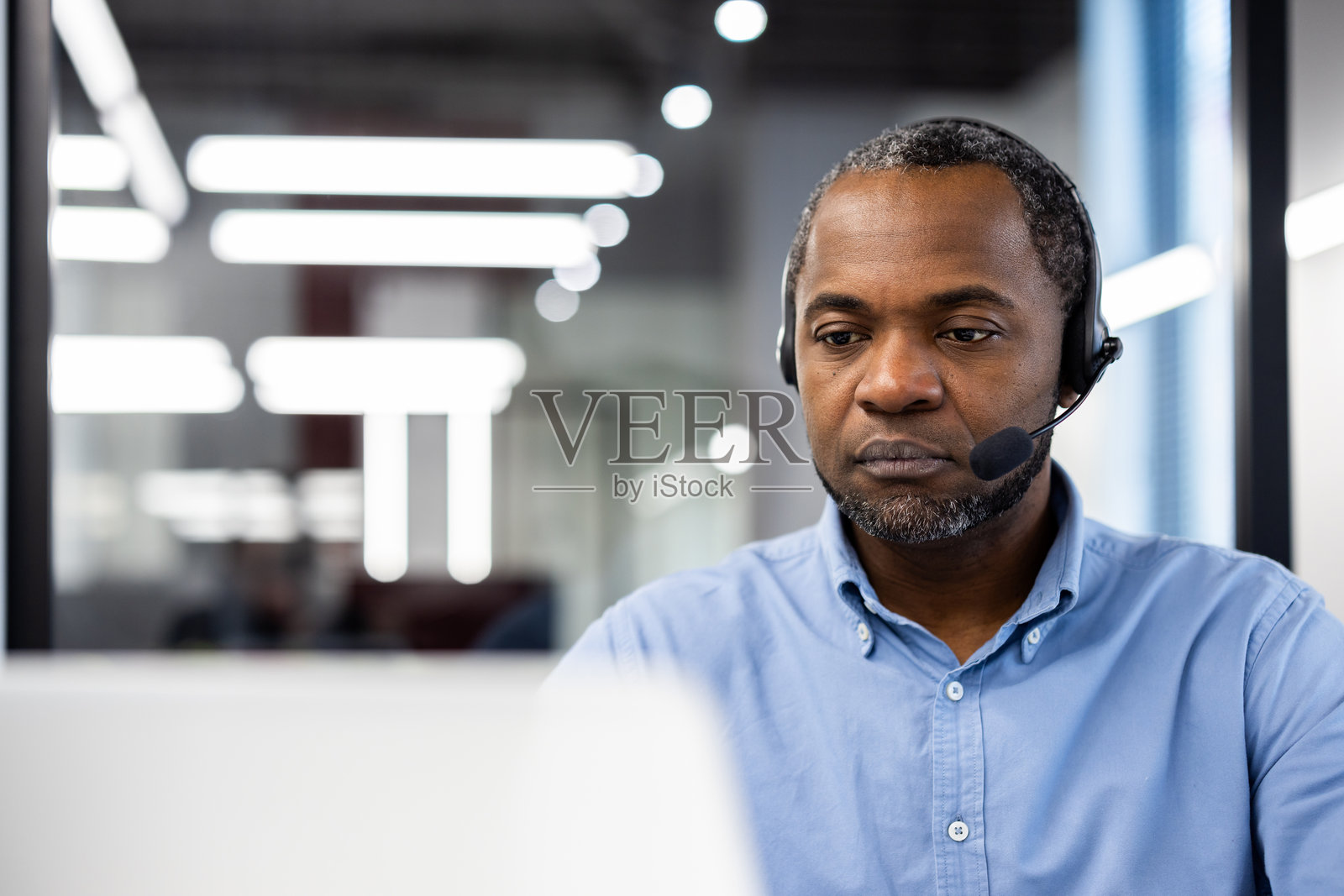 一位非裔美国成熟男商人在现代办公室中佩戴耳机工作,展现出专业和专注的企业环境,同时参与虚拟会议。照片摄影图片
