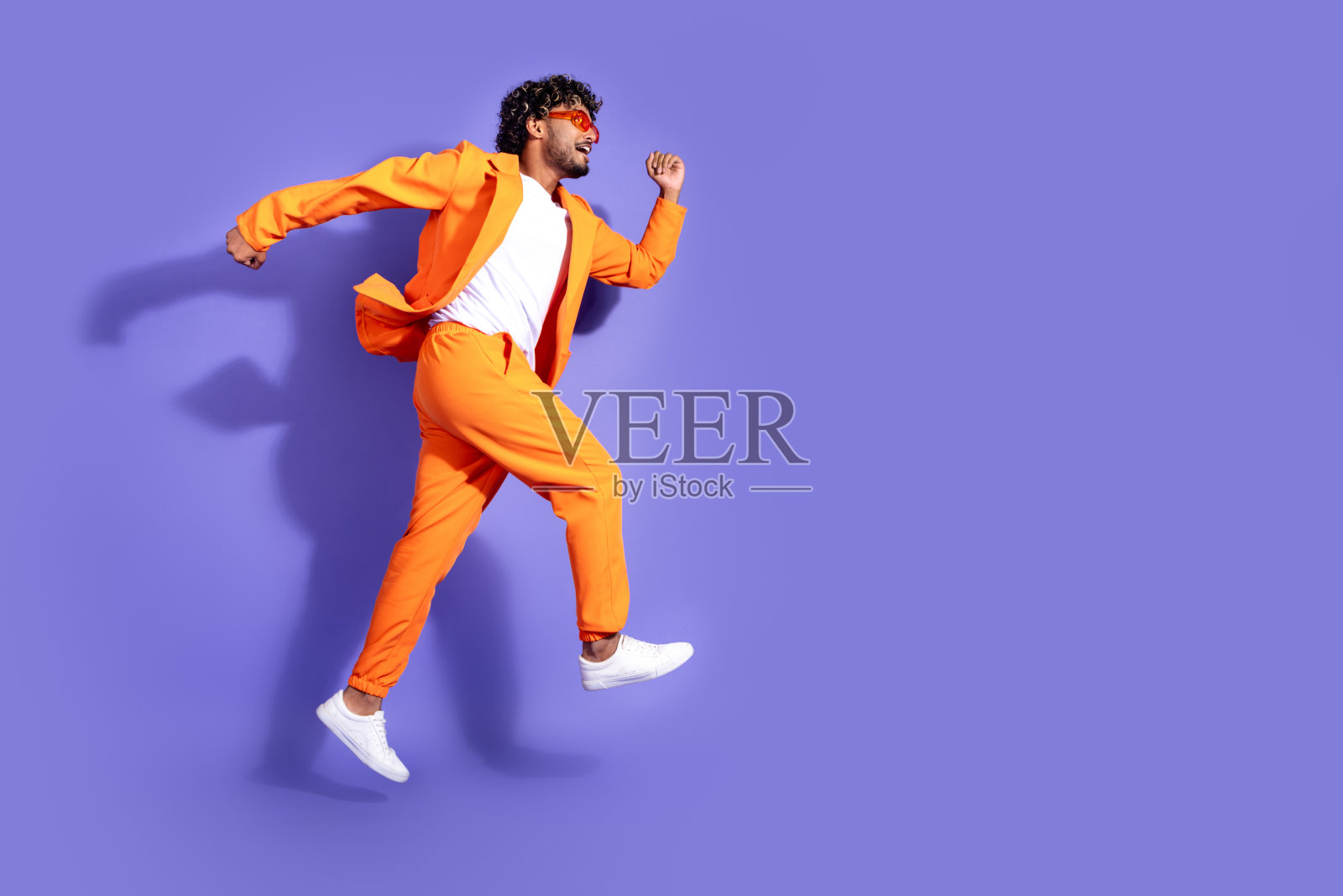 穿着橙色夹克的时尚酷男在紫色背景下高高跳起,展现出活力四射的姿态。照片摄影图片