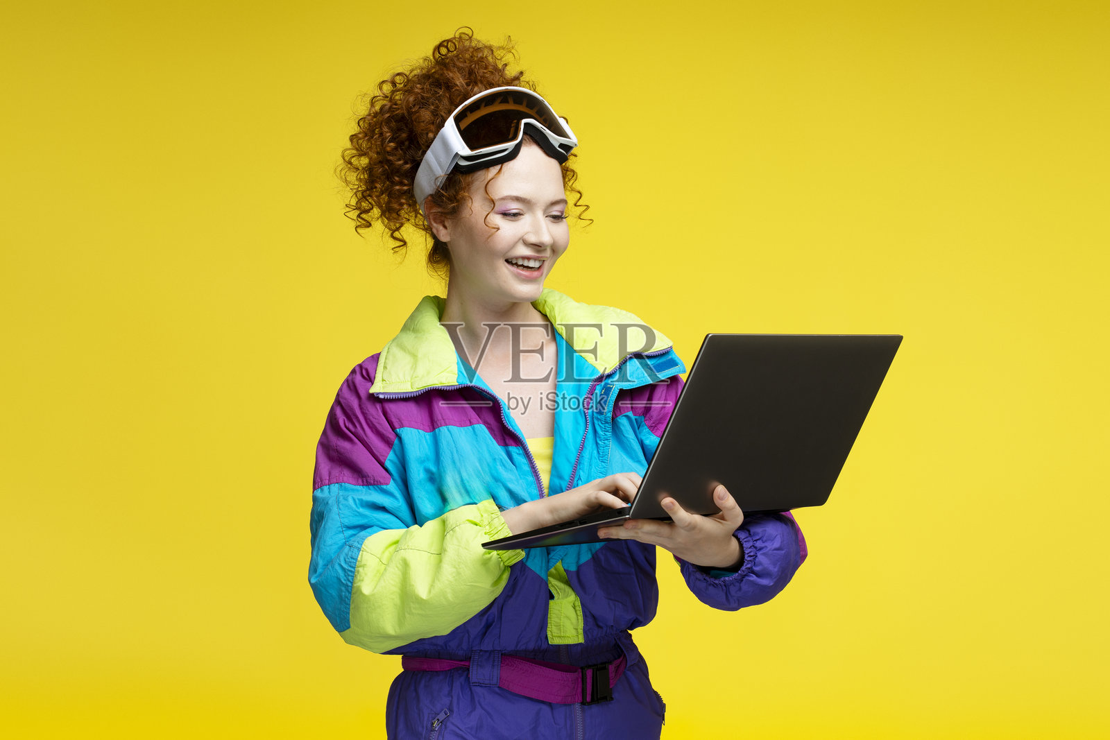 微笑的女性穿着滑雪服和护目镜,在黄色背景下使用笔记本电脑照片摄影图片