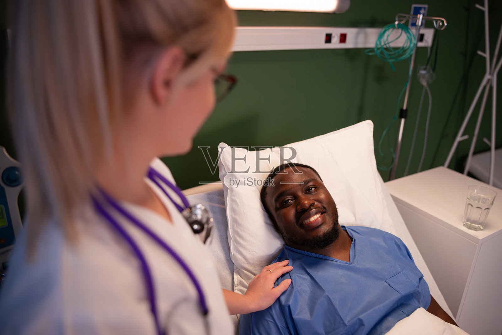 快乐的金发女医生在医院病房里微笑着站在非洲患者身旁,支持患者在经历严重事故后恢复。照片摄影图片