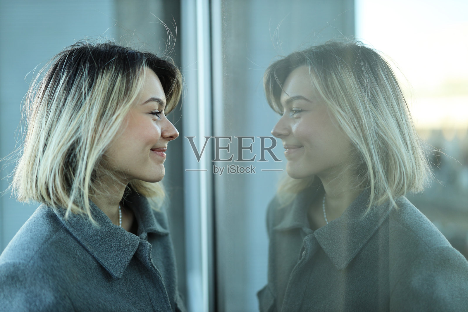 可爱的白人现代女孩看着窗玻璃中的倒影,微笑着,对自己的外貌感到满意。概念:年轻女性的城市生活场景照片摄影图片