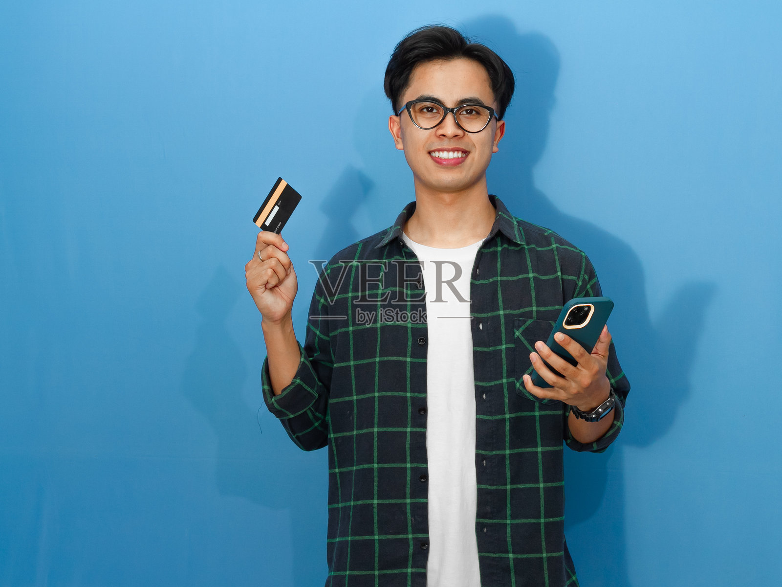 一位年轻的亚洲男子穿着休闲衬衫,手持银行卡和手机,面带自信微笑的表情。照片摄影图片