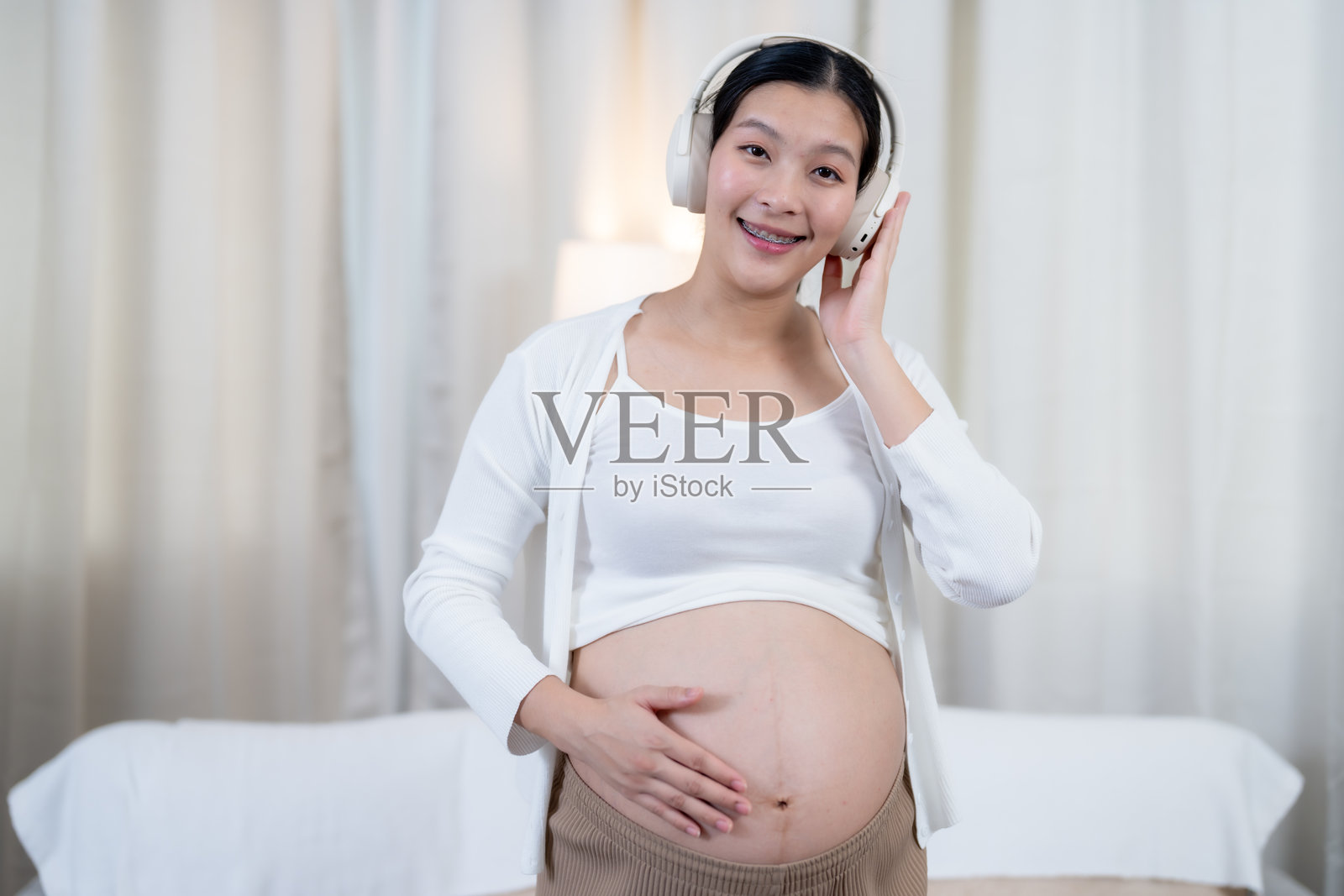 微笑的亚洲孕妇戴着耳机听音乐,手托着肚子。柔和的灯光和宁静的氛围反映出与未出生孩子的快乐和联系,拥抱母亲身份。照片摄影图片