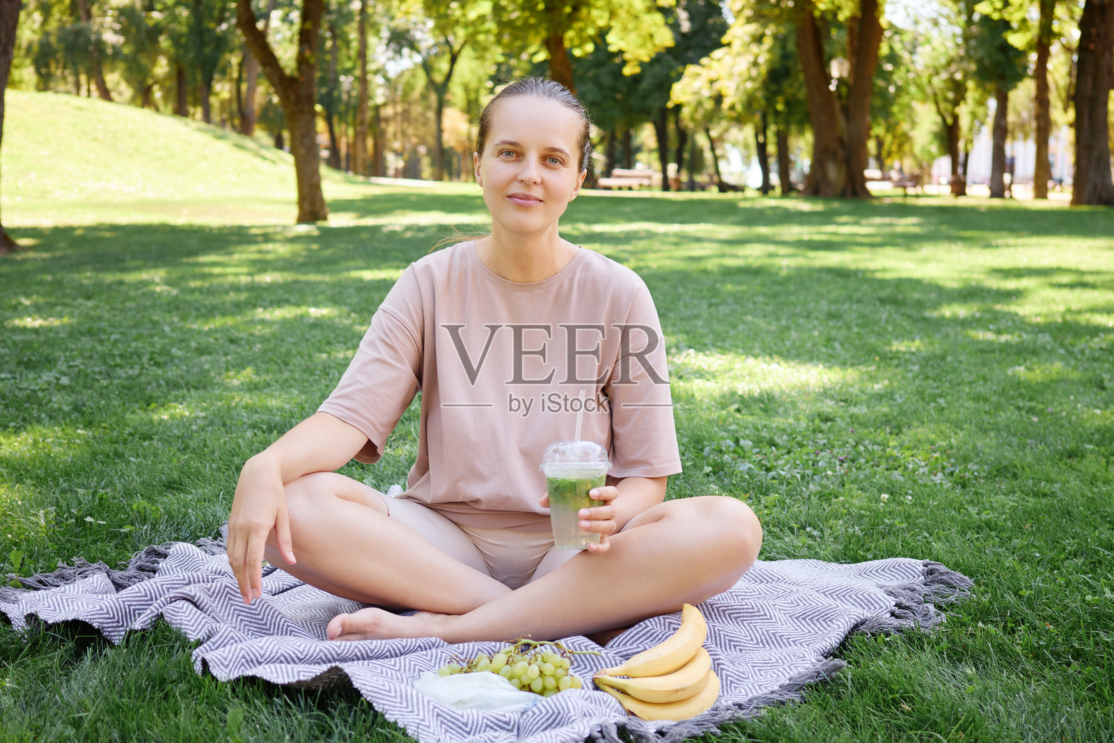 美丽的年轻女性在花园里野餐,坐在绿草地上的毯子上,享用水果和饮用柠檬水或鸡尾酒,享受夏日的休闲时光。照片摄影图片
