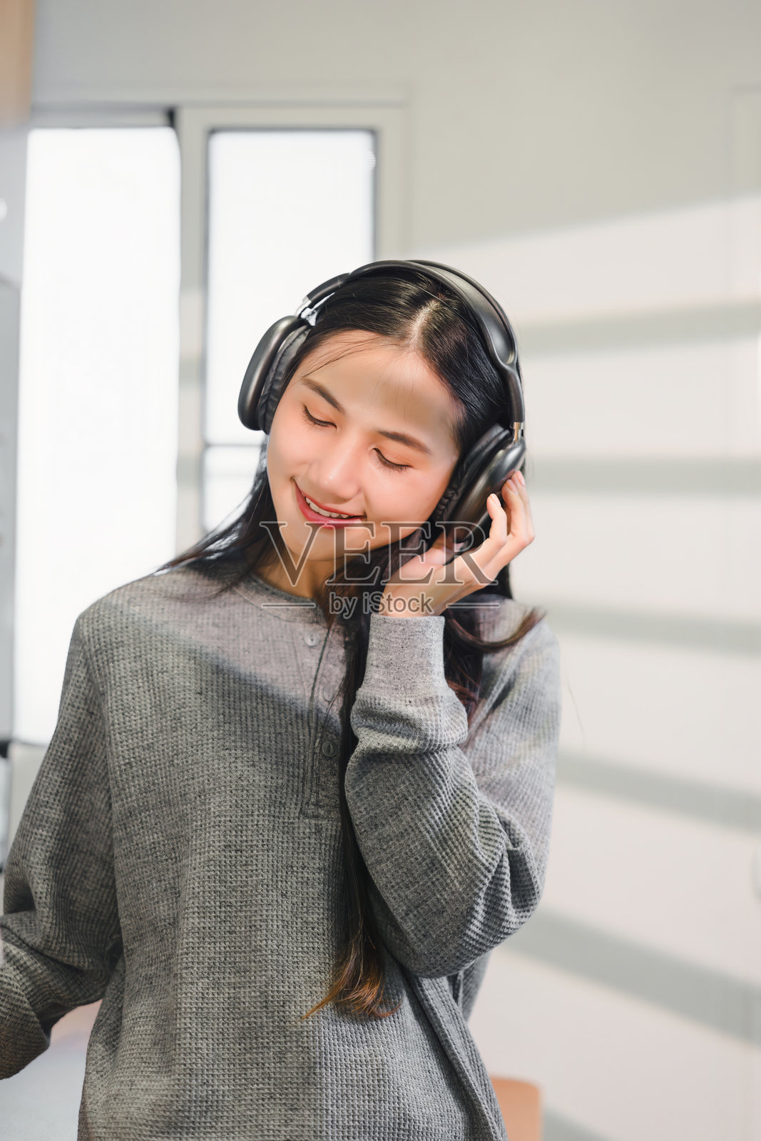 年轻的亚洲女性在室内微笑着享受音乐,佩戴着耳机。柔和的光线营造出温暖的氛围,增强了她的愉悦体验。照片摄影图片