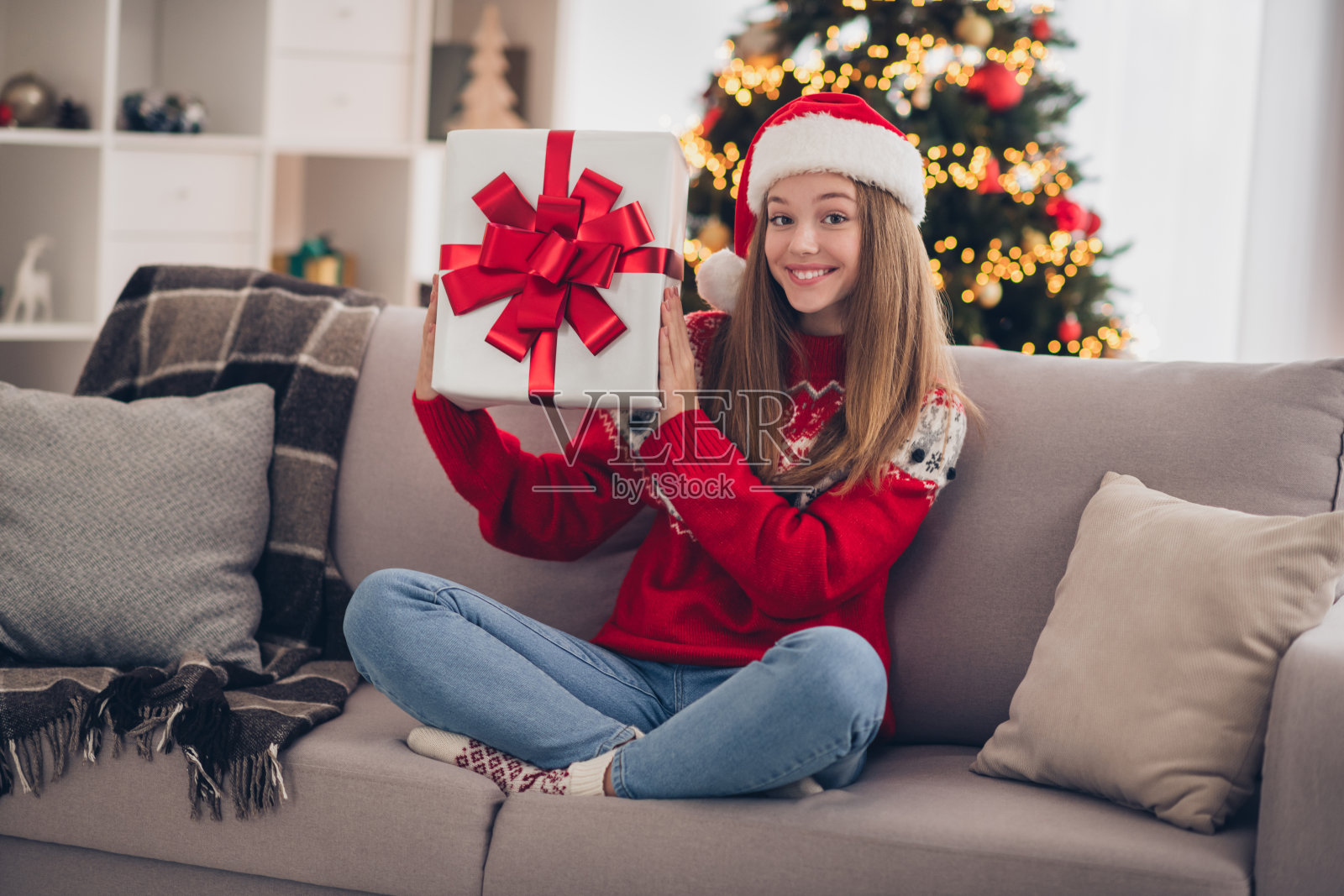 全身肖像,积极甜美的人坐在舒适的沙发上,双手捧着礼品盒,圣诞节的室内环境。照片摄影图片