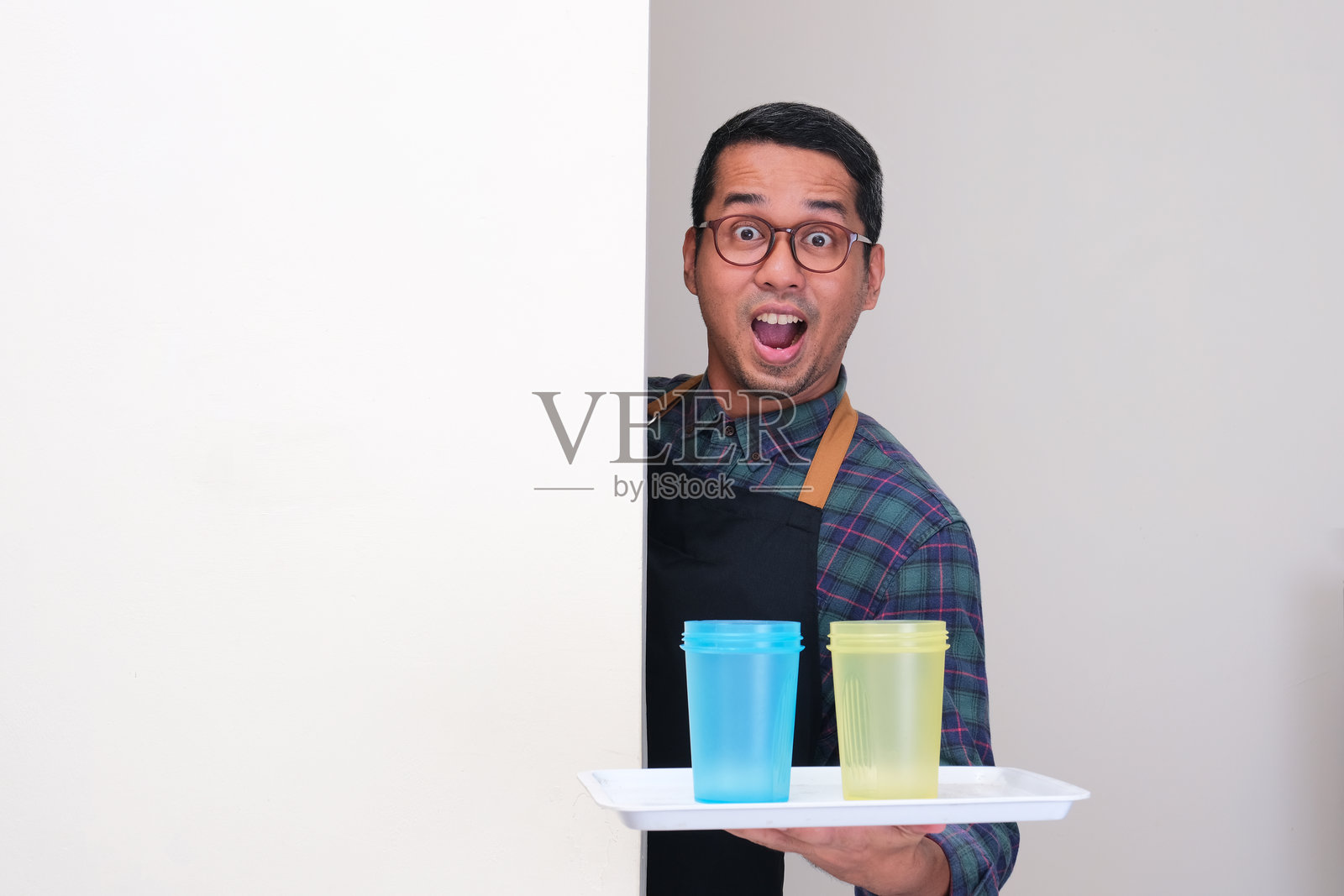 一个穿着围裙的男人在墙后探头,脸上露出惊讶的表情,手里托着两个饮料瓶。照片摄影图片