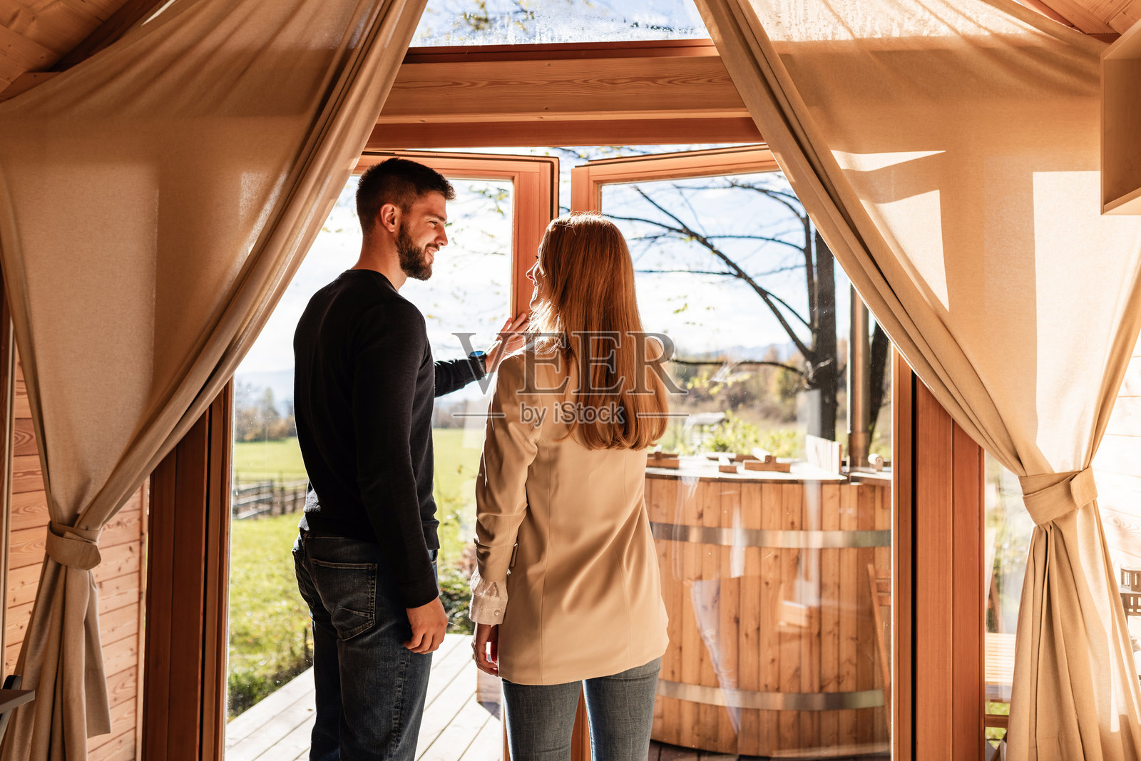 一对情侣在带全景窗户的豪华露营小屋里,露台上有一个木制热水浴缸。照片摄影图片