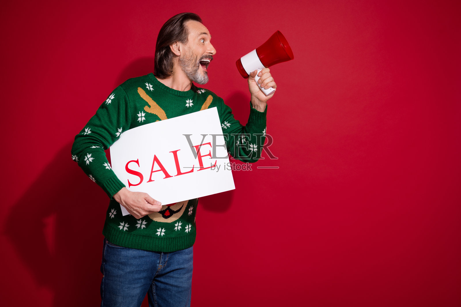 兴奋搞笑的人手持扬声器和纸质促销标牌,传达消息,背景为空旷的红色。照片摄影图片
