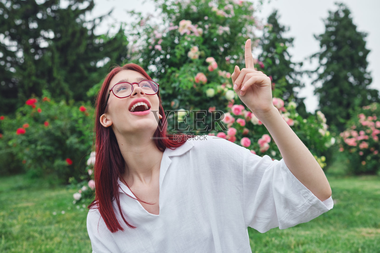 红发戴眼镜的少女在盛开的花园里,抬头指着上方,露出惊讶的表情。照片摄影图片