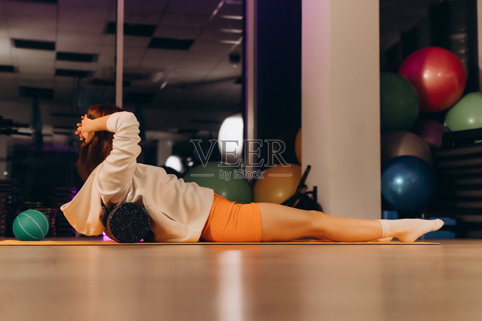 美丽的年轻白人女性运动员使用泡沫滚轮按摩器放松,拉伸肌肉和缓解背痛。女孩穿着绿色运动鞋、T恤和紧身裤在健身房里。照片摄影图片