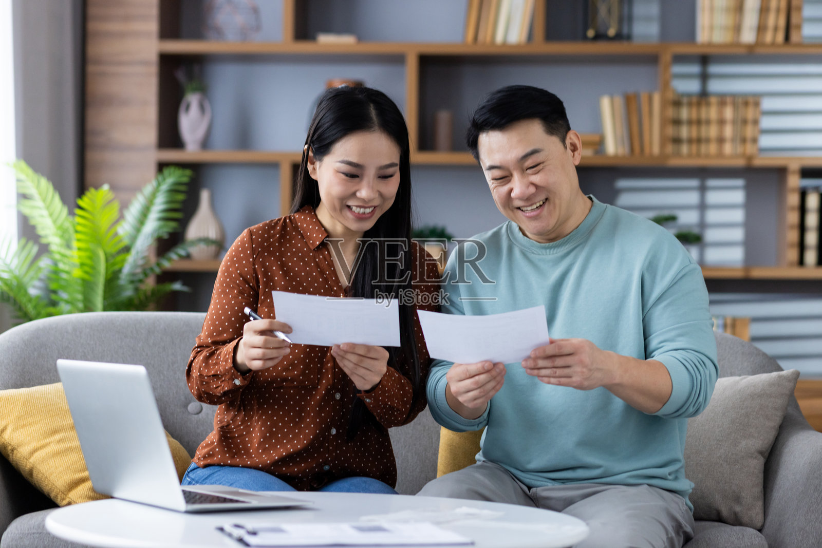 快乐的亚洲夫妇在家中使用笔记本电脑审阅文件,展现团队合作、财务管理和协作,周围是温馨舒适的客厅环境,充满温暖和支持的氛围。照片摄影图片