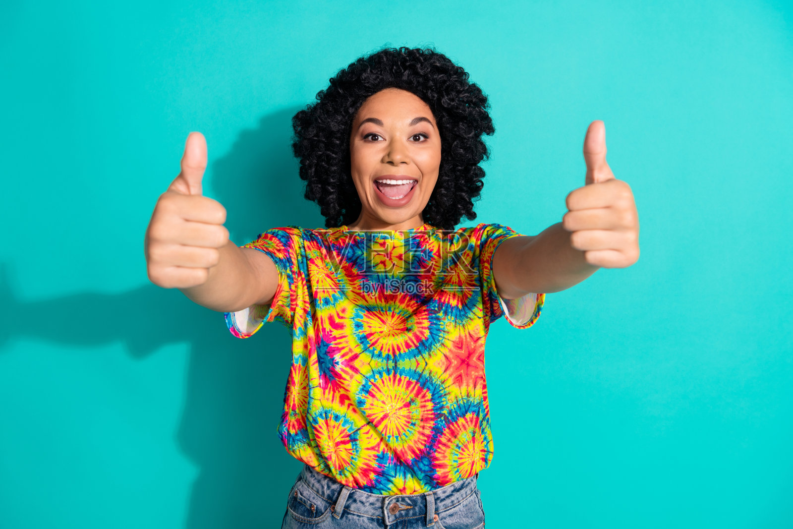 穿着时尚印花衣服的可爱满意女性在青色背景前竖起大拇指。照片摄影图片