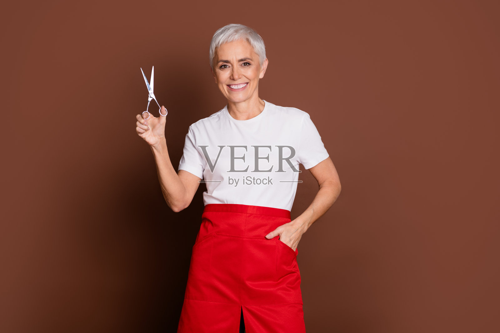 美丽的资深女性专业发型师在美容院里手持剪刀,穿着制服,背景为棕色。照片摄影图片