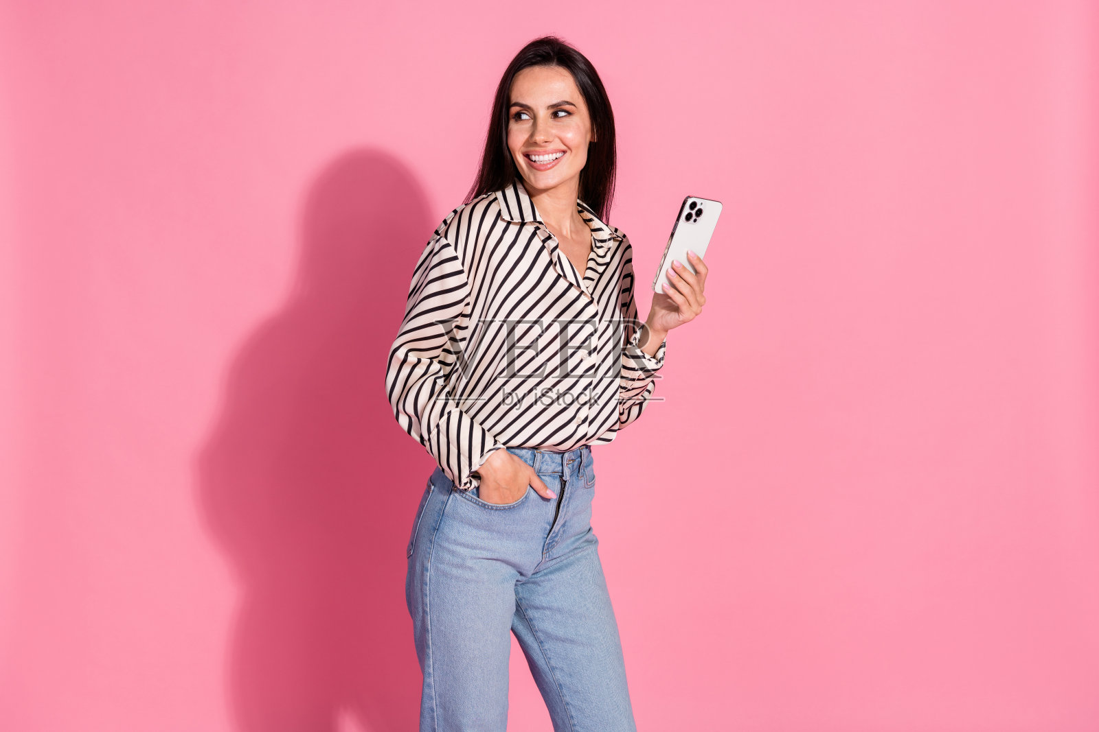 年轻女性,棕色头发,穿着休闲条纹衬衫和牛仔裤,手持智能手机,背景为粉色。照片摄影图片