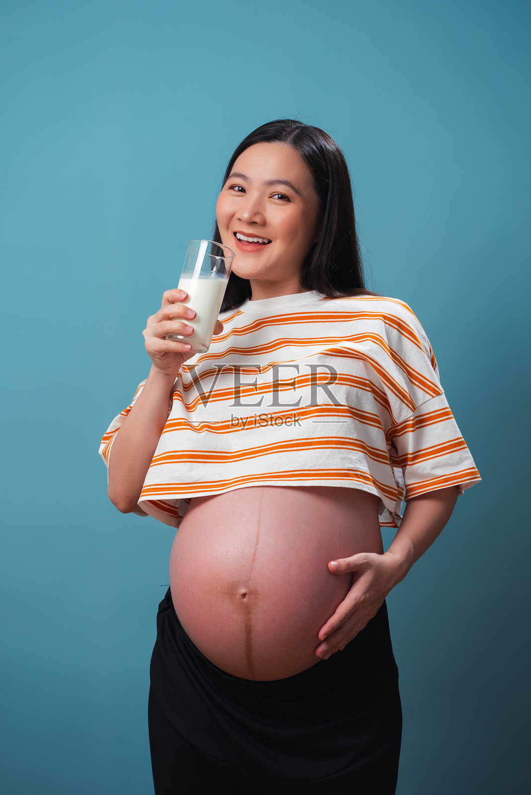 亚洲孕妇手握牛奶杯,手抚肚子,面向镜头,背景为蓝色。照片摄影图片