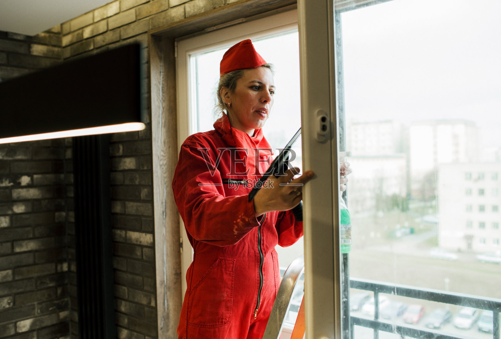 一位穿着红色连体衣的女性正在用刮水器和窗户清洁剂清洁窗户。照片摄影图片