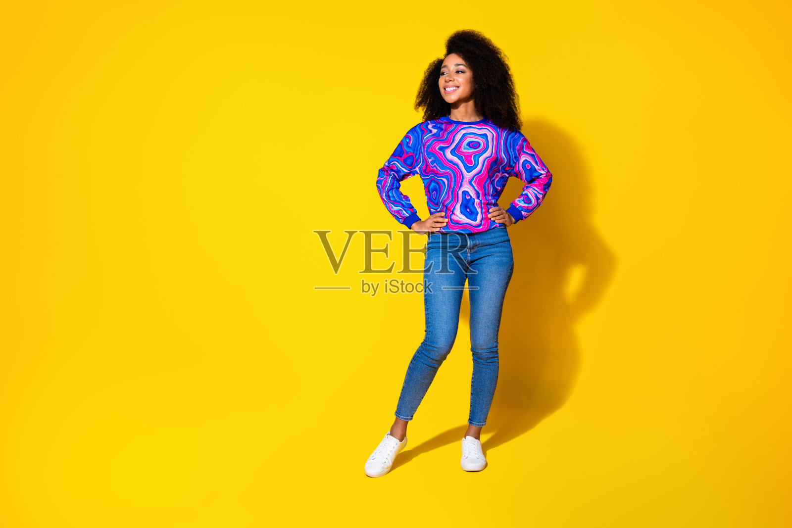 全身照,吸引人的年轻女性穿着时尚的彩色蓝色服装,摆姿势看向空旷的空间,背景为黄色。照片摄影图片