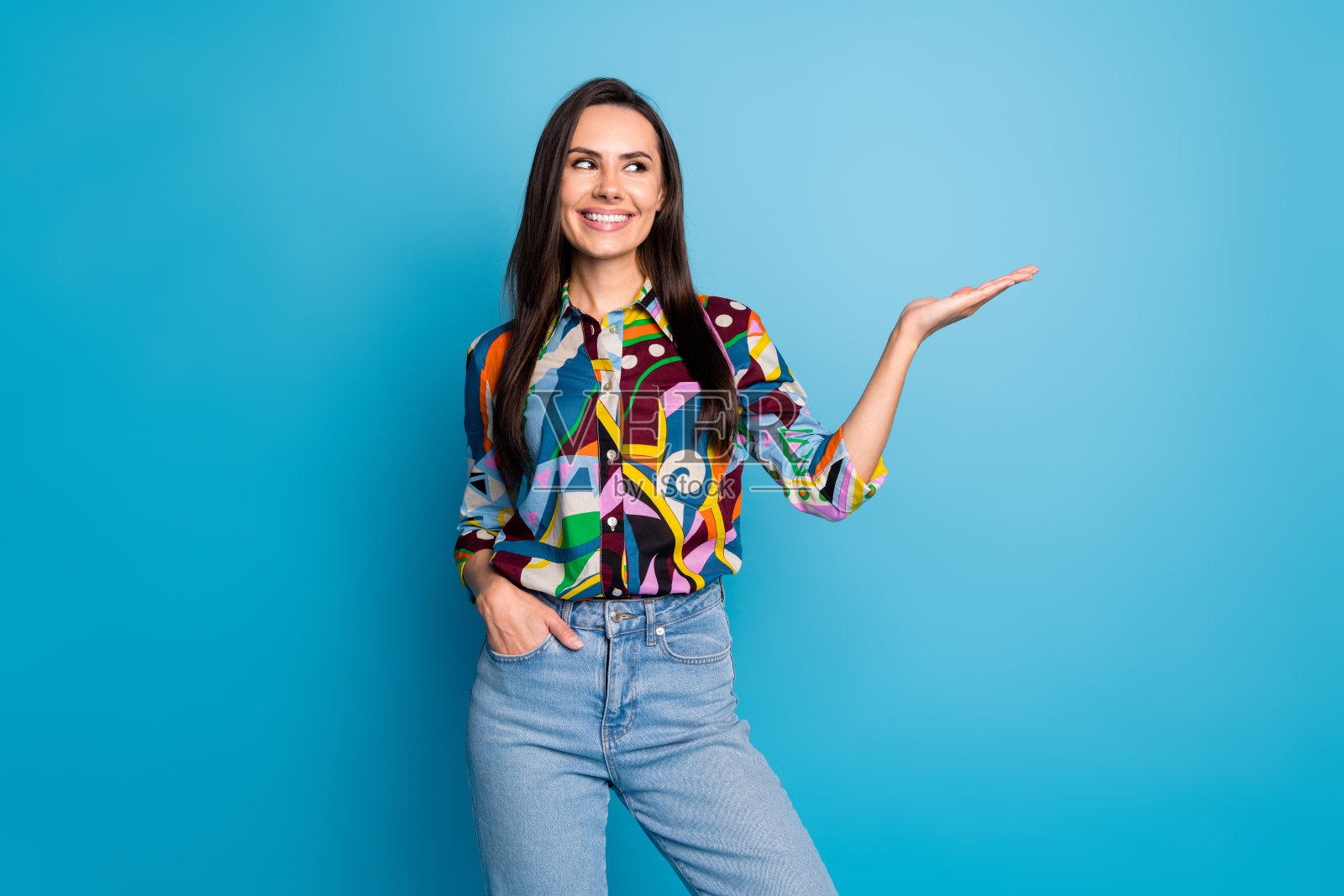时尚的年轻女性在蓝色背景下微笑,穿着色彩鲜艳的印花衣服,手中指向空白空间。照片摄影图片
