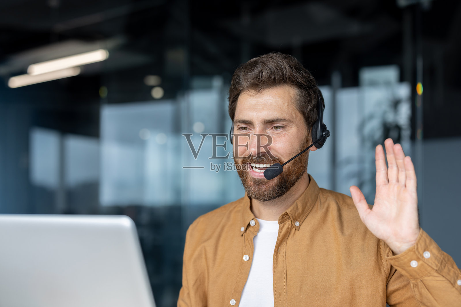 自信的男士在现代办公室进行在线视频通话,佩戴耳机,挥手交流。照片摄影图片