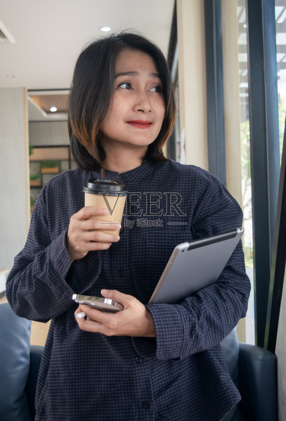 亚洲女性在咖啡馆里站着,手里拿着咖啡杯和平板电脑,同时用手机,望向窗外,享受放松的假期生活方式。照片摄影图片