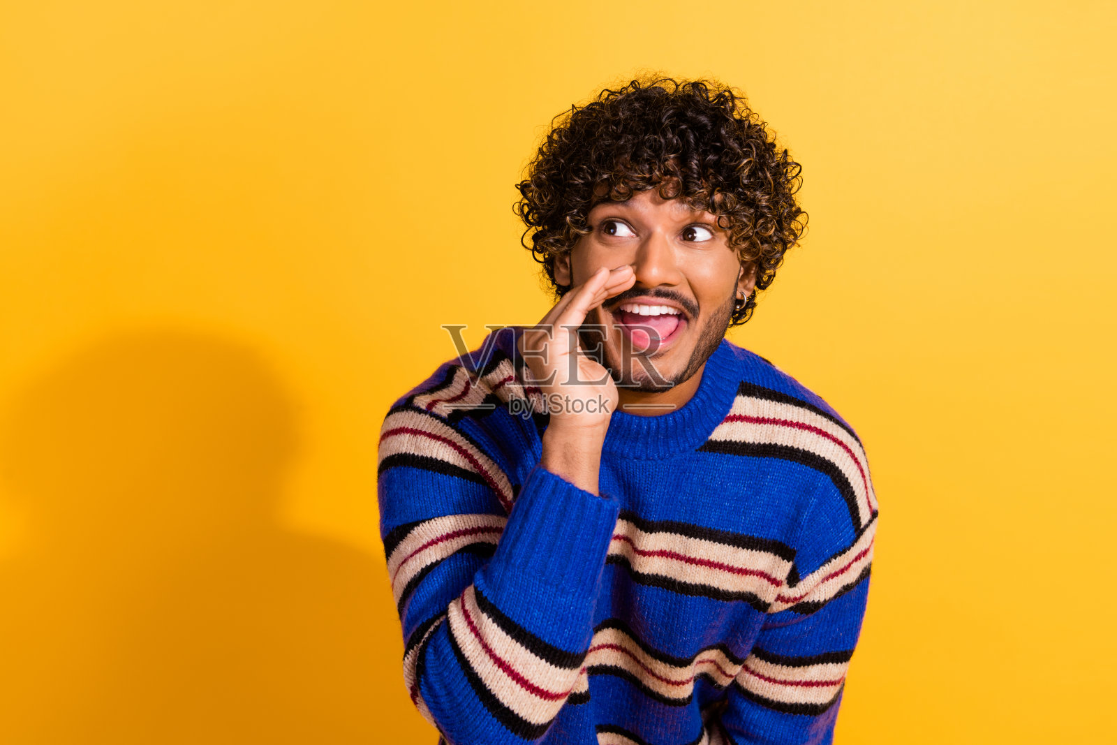 穿着时尚蓝色条纹衣服的年轻帅哥捂住嘴巴尖叫,望向空旷的空间,背景为黄色。照片摄影图片