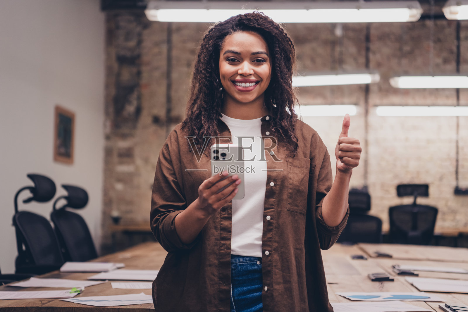 成功的年轻女商人在现代商务中心办公室内举起手机竖起大拇指,穿着衬衫。照片摄影图片