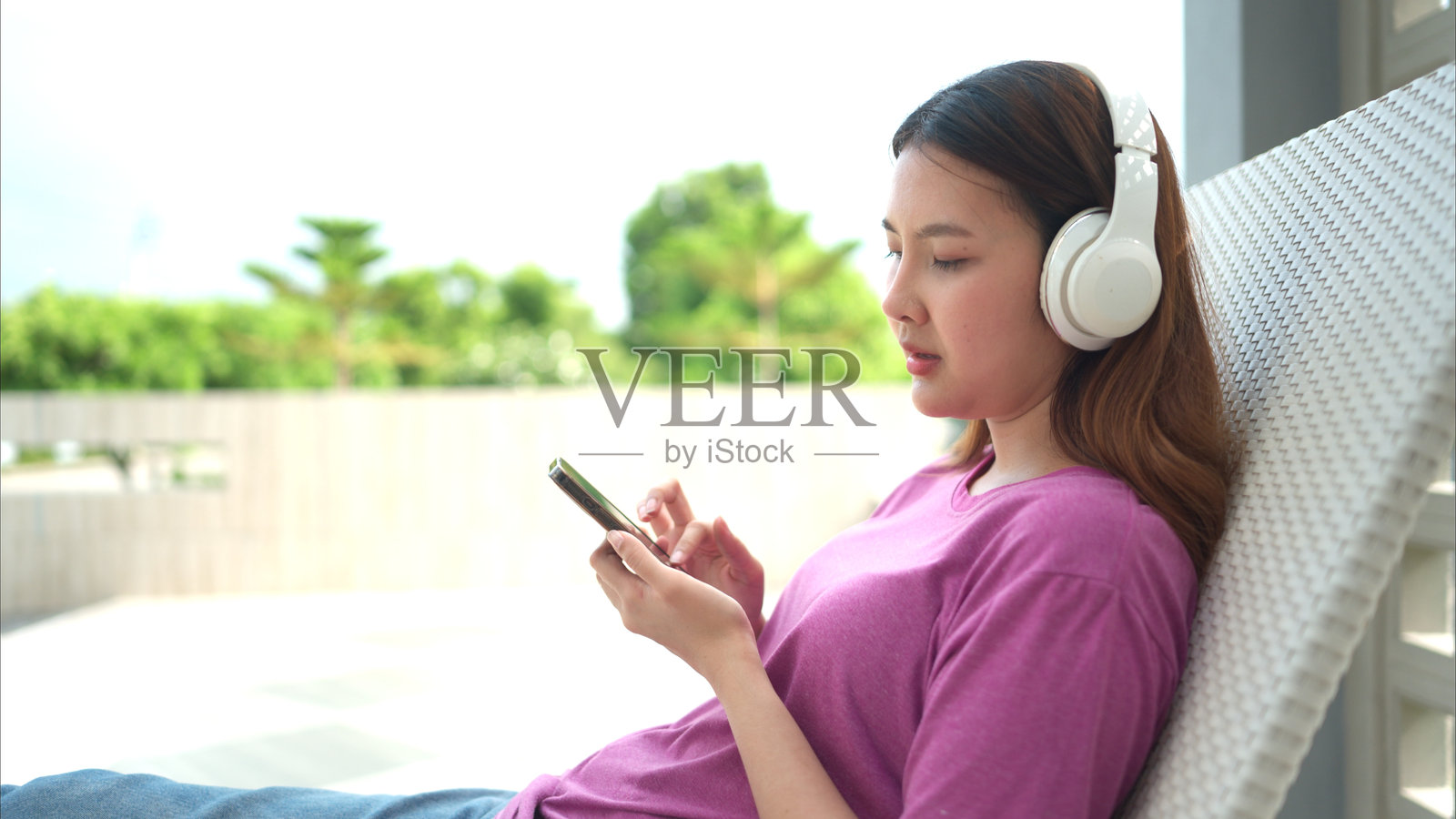 亚洲年轻女性愉快地坐在躺椅上,戴着耳机听音乐。照片摄影图片