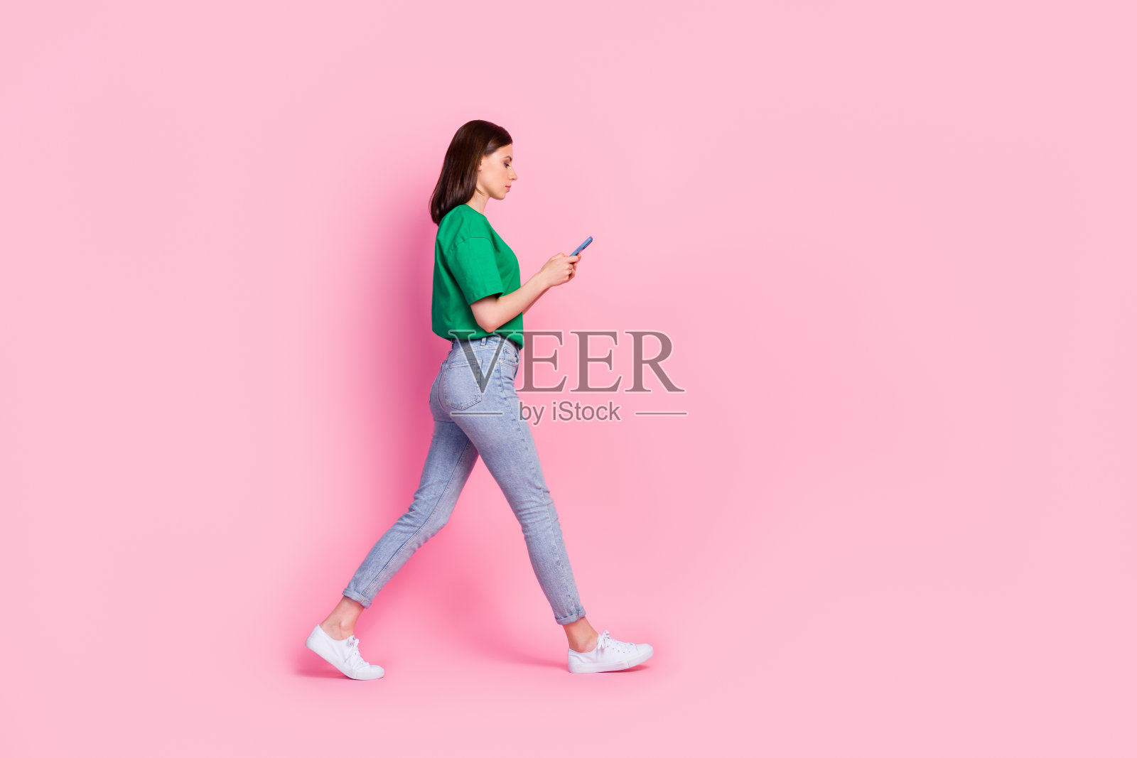 全身照,漂亮的年轻女孩穿着时尚的绿色衣服,走路时手持设备聊天并阅读新闻,背景为粉色。照片摄影图片