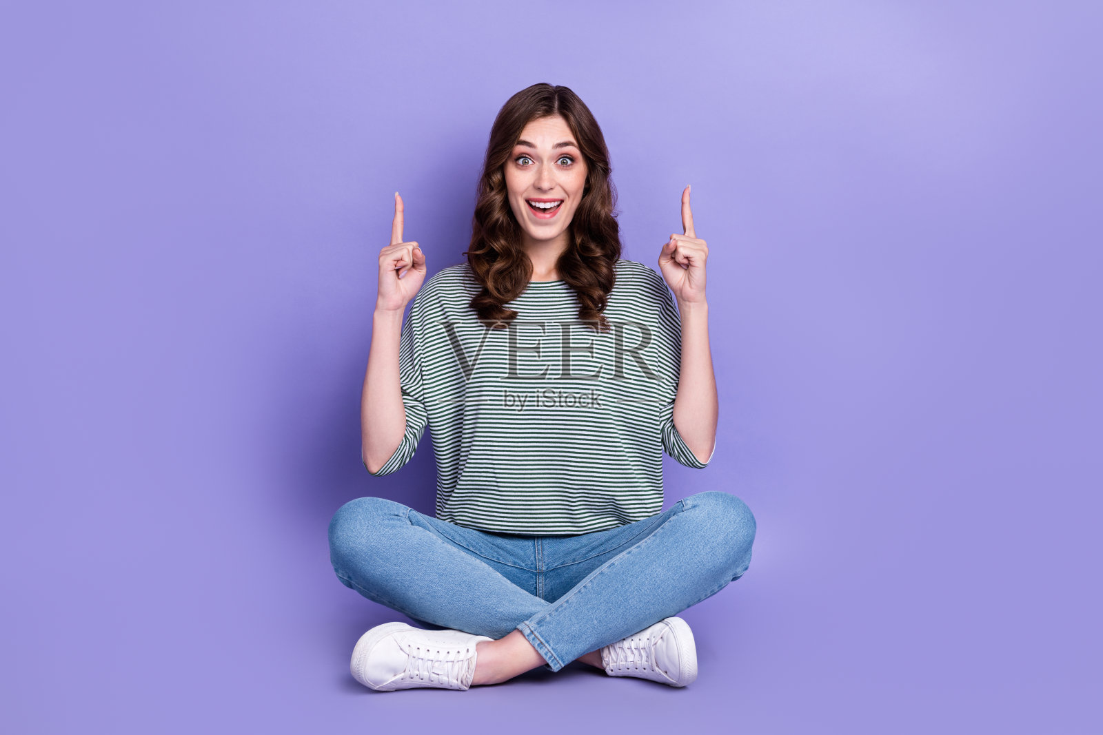 惊喜而快乐的女士坐在空旷的空间中,双臂伸展,展示低价时尚服装,背景为紫色。照片摄影图片