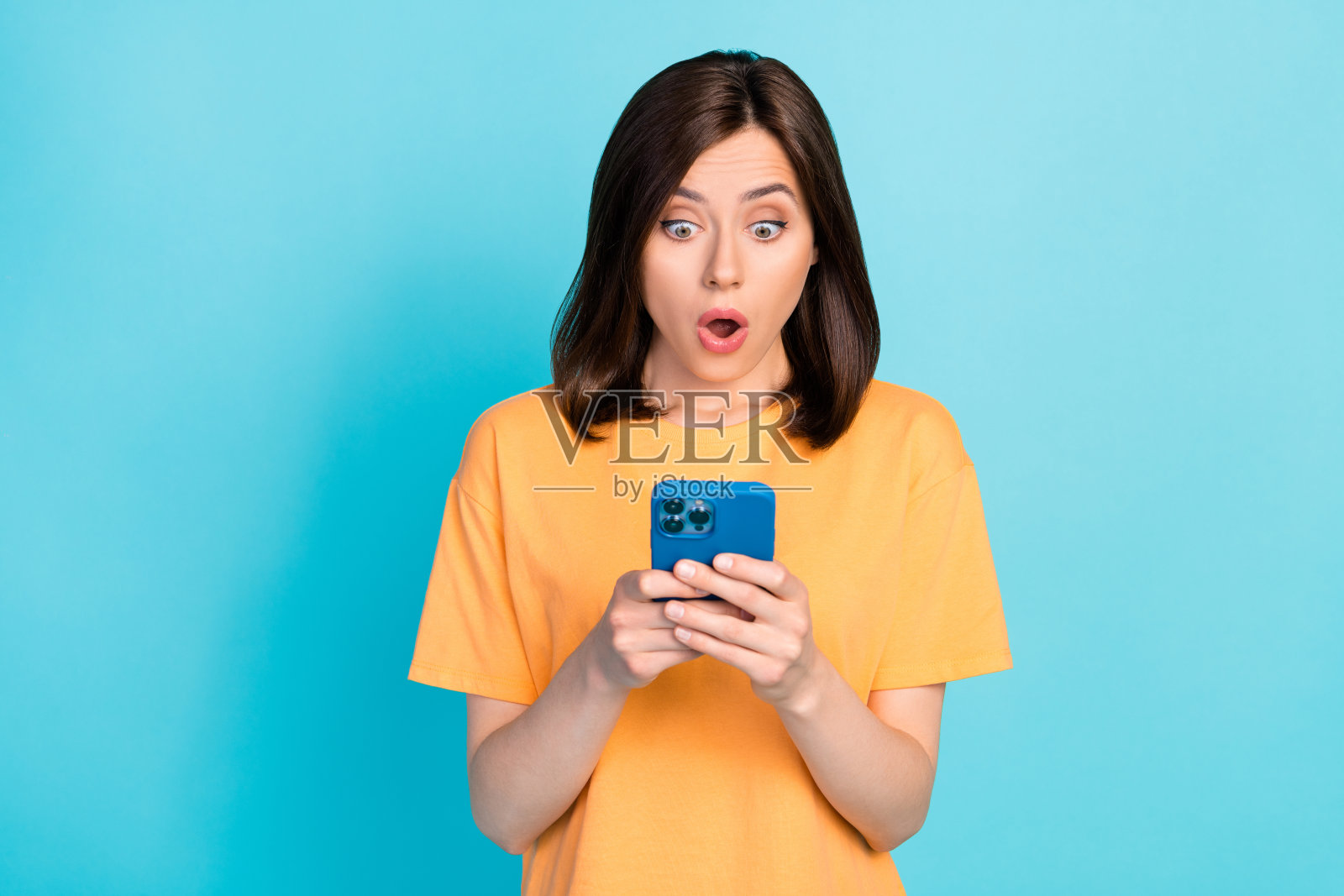 一位惊讶的漂亮女孩张嘴使用电话打字,背景为蓝色。照片摄影图片