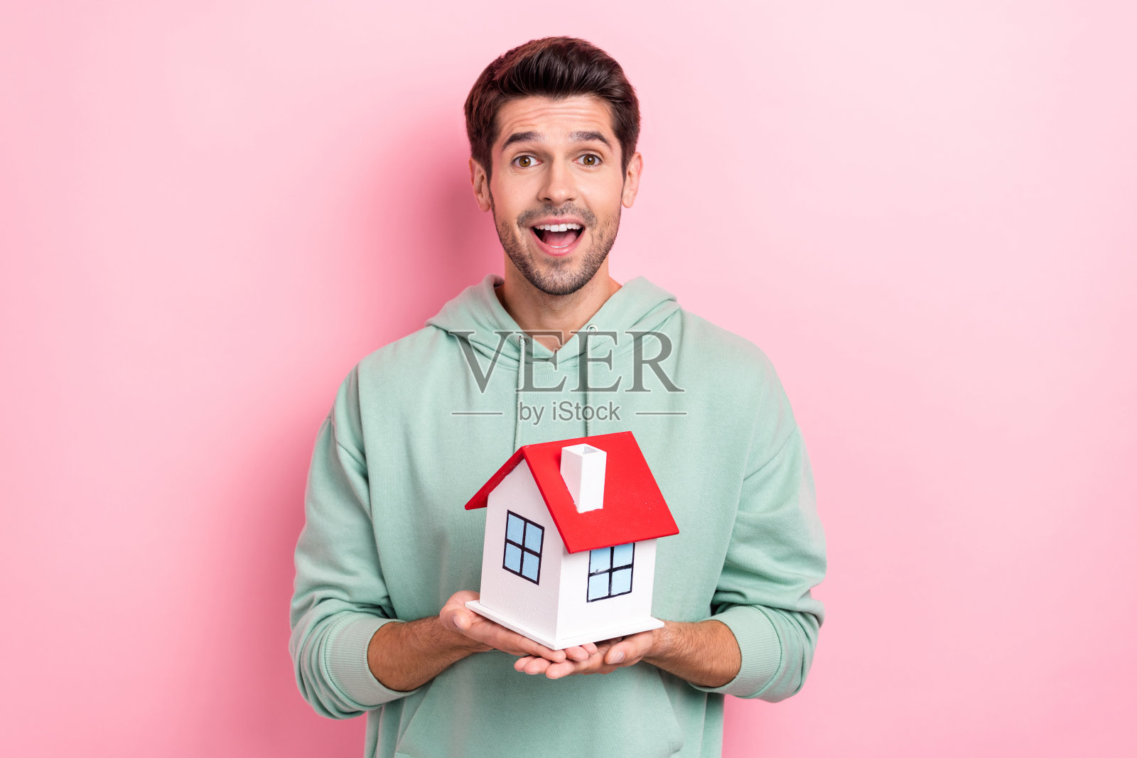 惊讶而快乐的男人手握小房子,张嘴微笑,背景为粉色。照片摄影图片