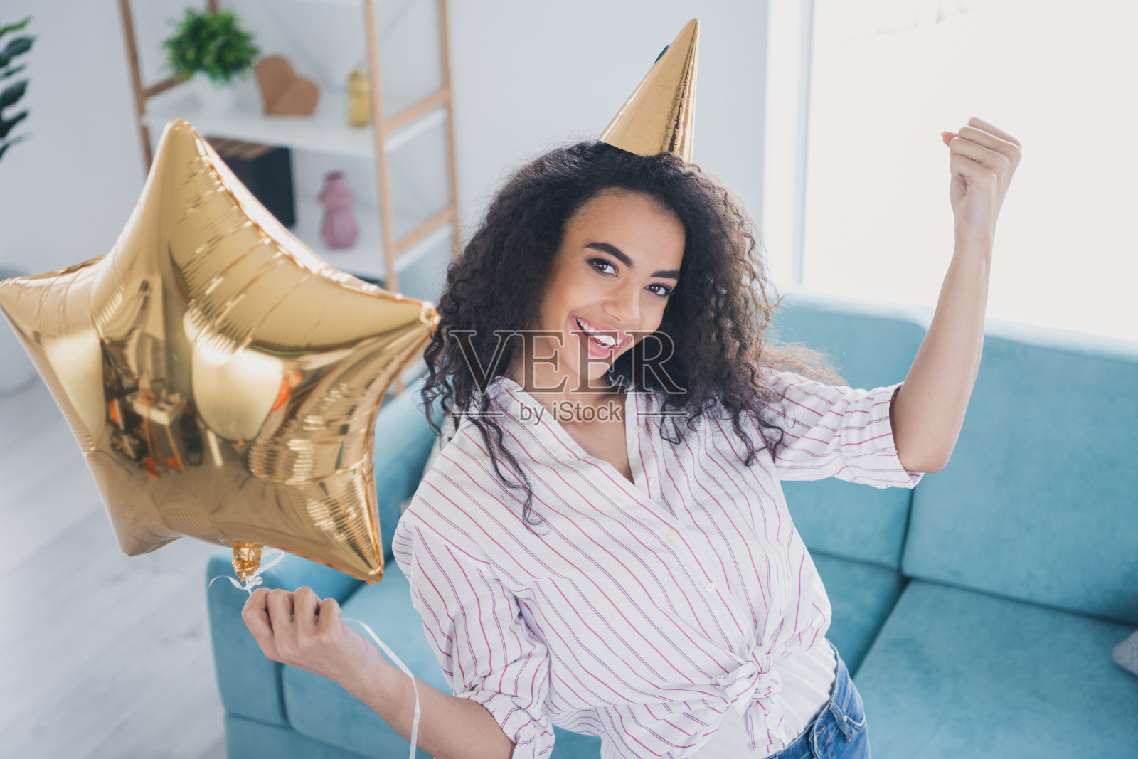 俯视角度的年轻女孩,举起拳头,戴着尖顶帽,手持星形气球,庆祝生日派对,室内场景。照片摄影图片