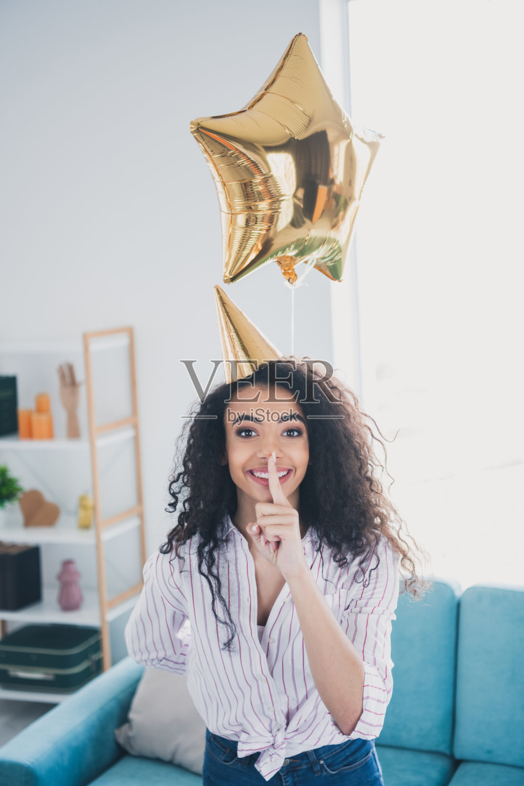 漂亮年轻女孩竖版肖像,做出嘘的手势,戴着尖顶帽,手持星形气球,生日派对场景,室内平面图。照片摄影图片